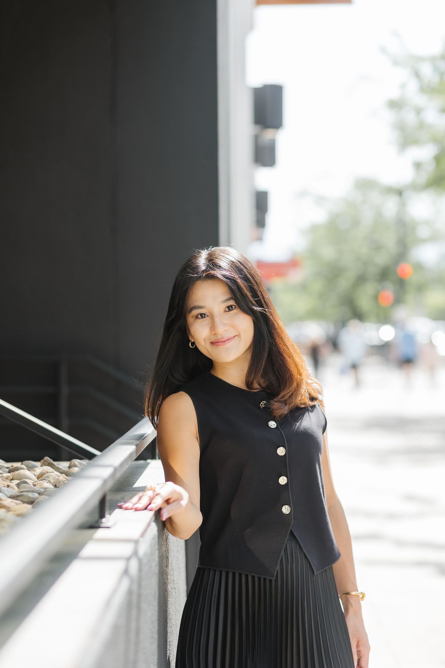 A person wearing a sleeveless black dress poses against a modern architectural backdrop with glass windows.