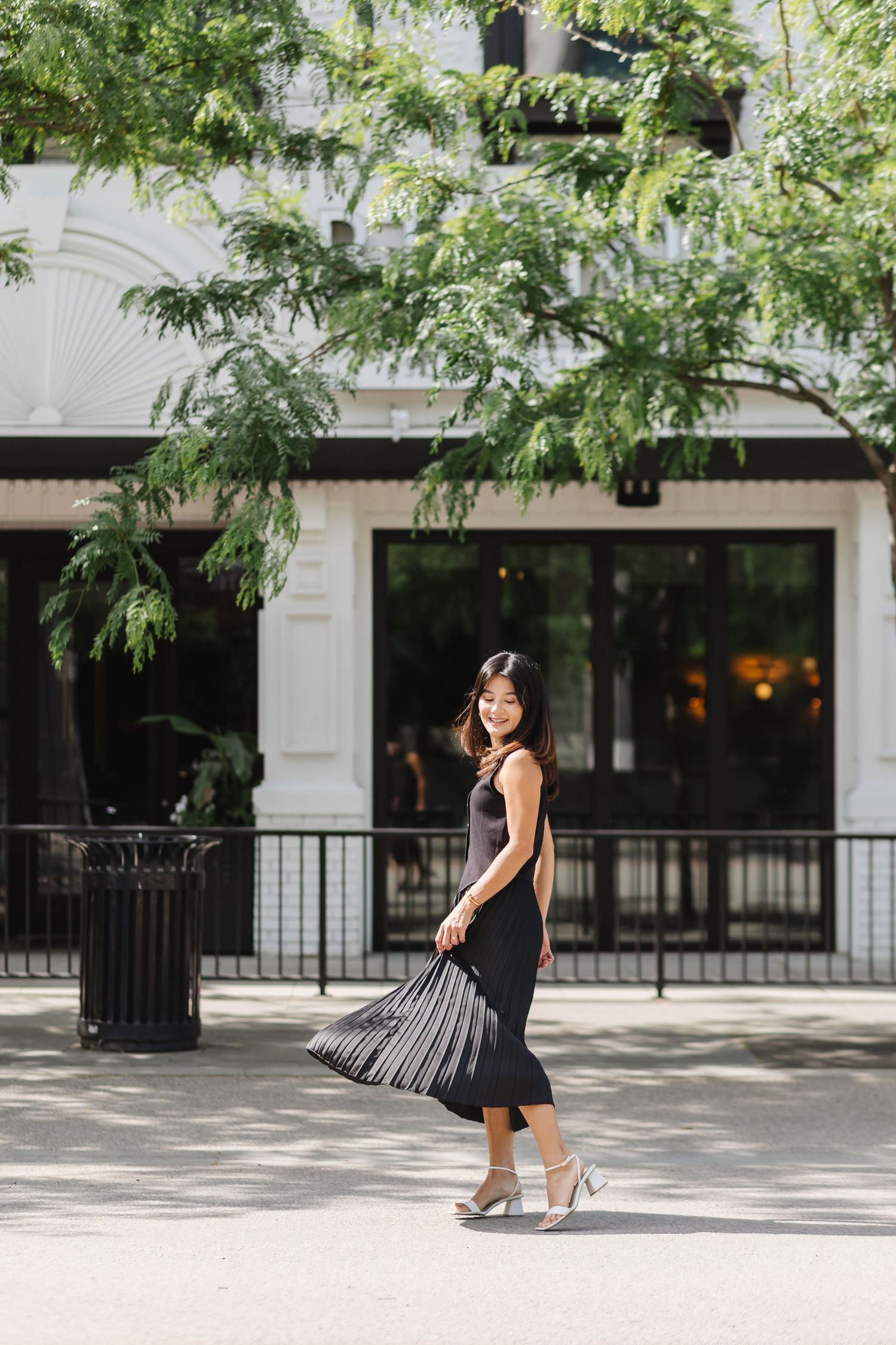 A person in a black dress performs a playful dance sequence against an architectural backdrop with overhanging trees.