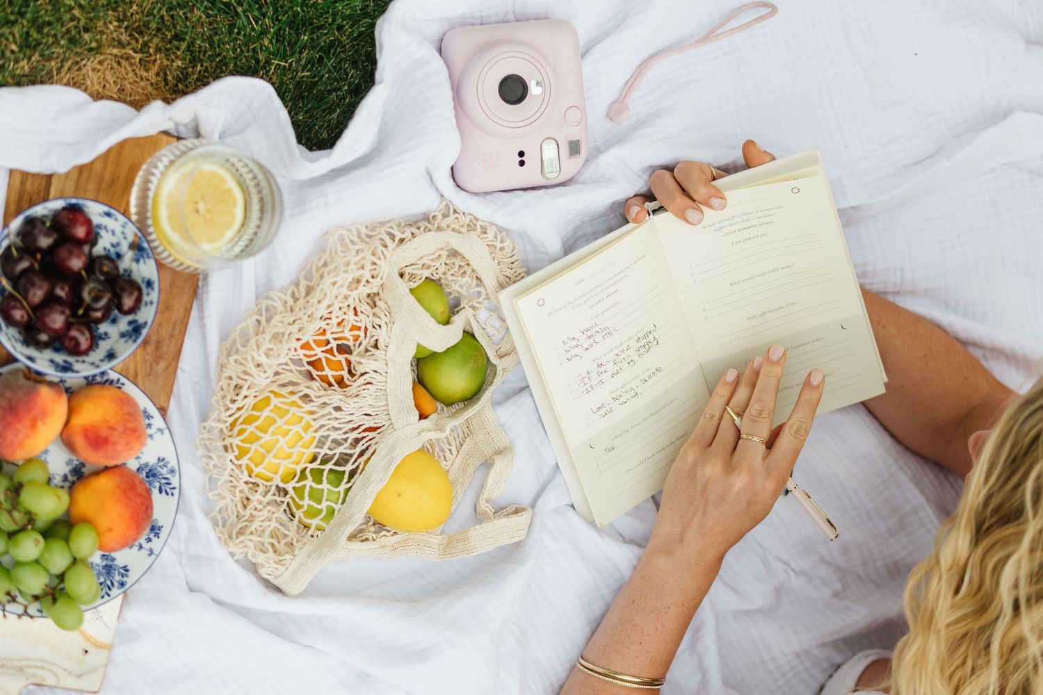 Overhead view of a picnic blanket with fresh fruit, notebook, and reusable produce bags in natural sunlight.