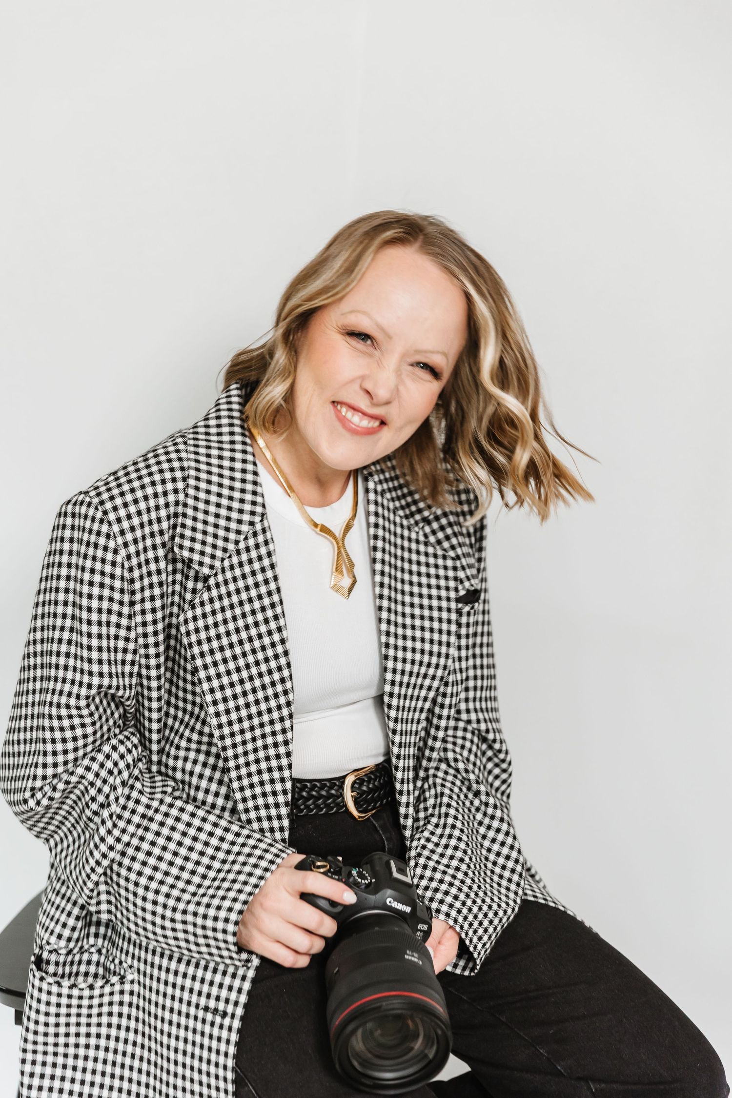Smiling blonde woman in checkered blazer holding a Canon DSLR camera, posing against a white background.