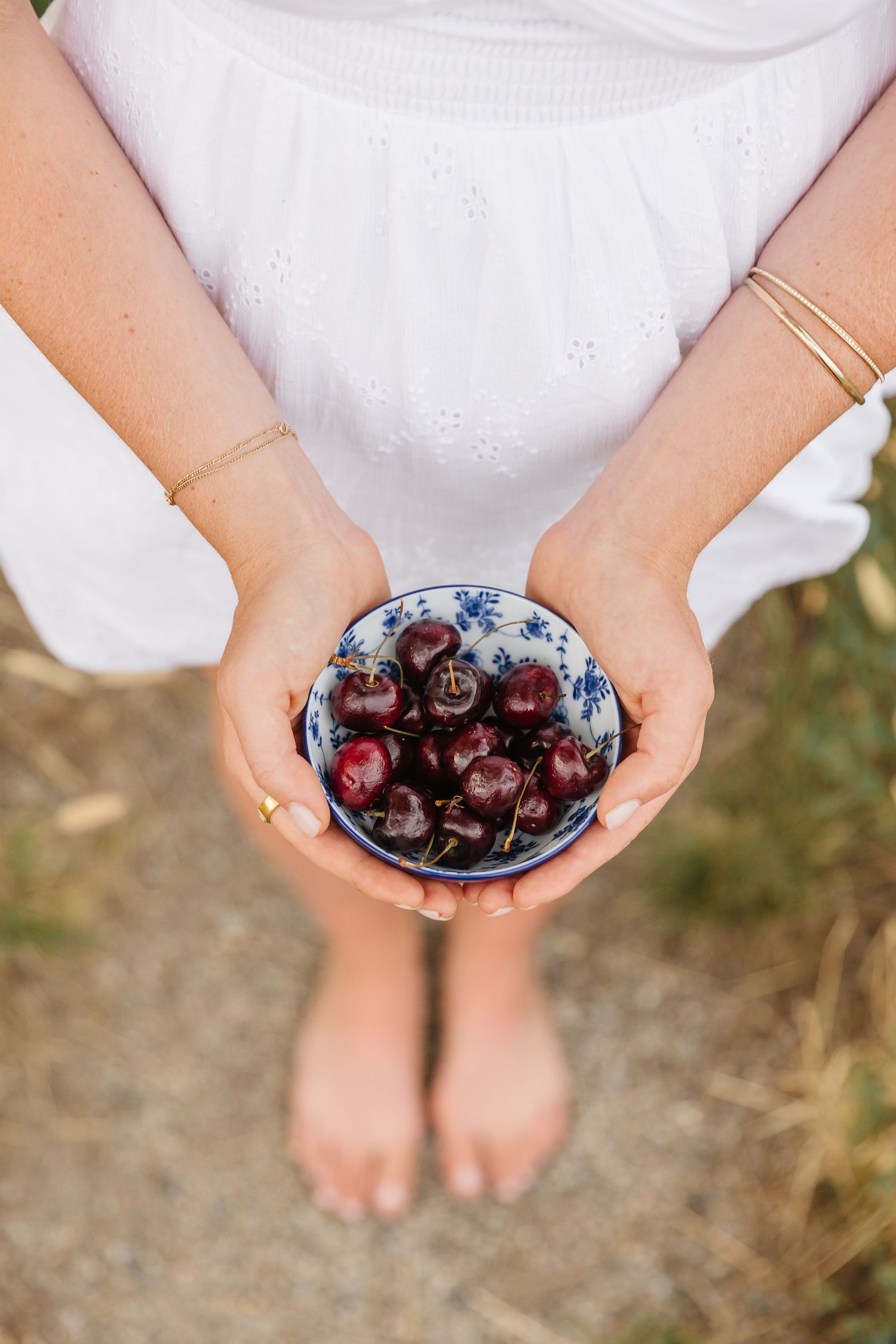 Hands holding a blue bowl filled with fresh dark cherries while wearing a white eyelet dress.