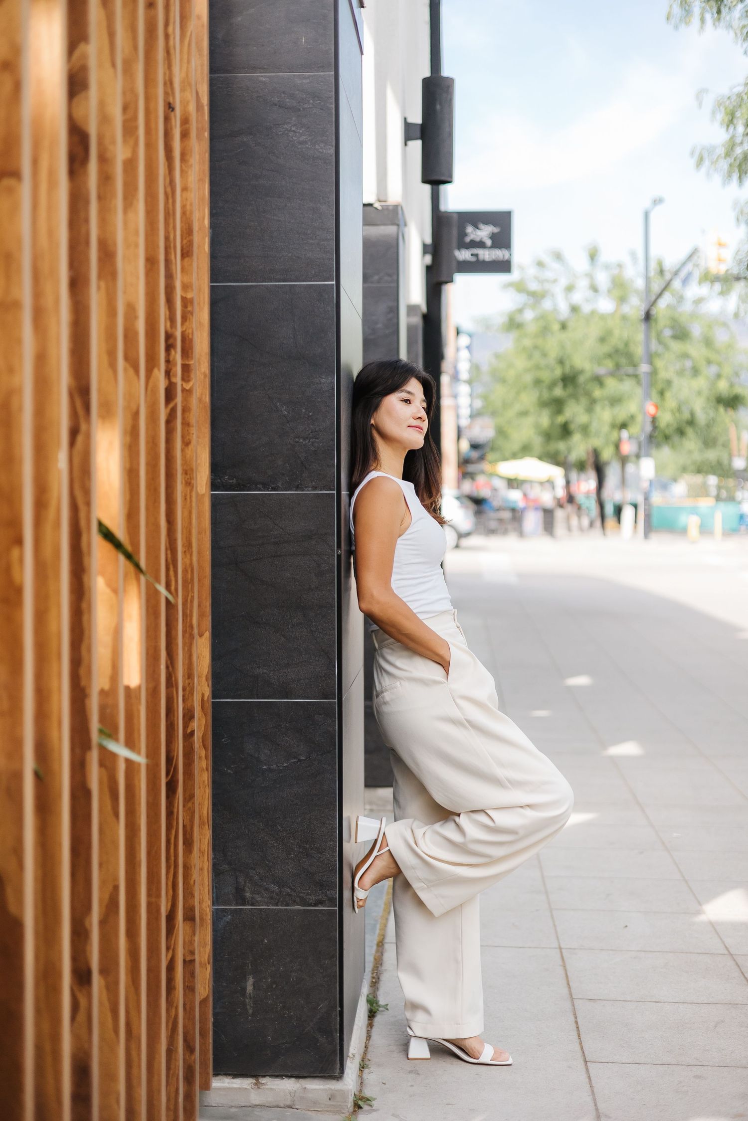 A casual summer street scene showing someone in a white tank top and cream pants leaning against a modern building exterior.