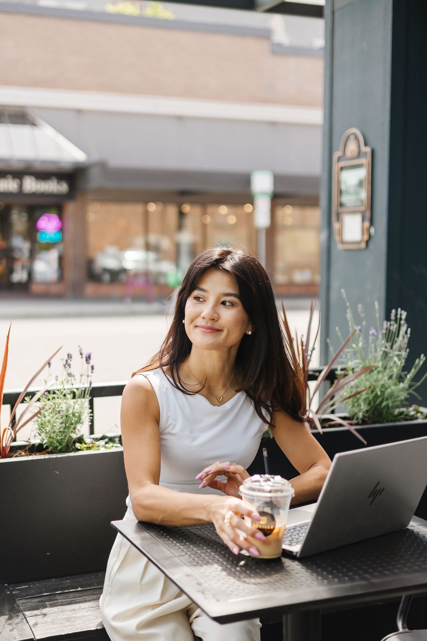Person working on laptop at outdoor cafe with plants and storefront in background.