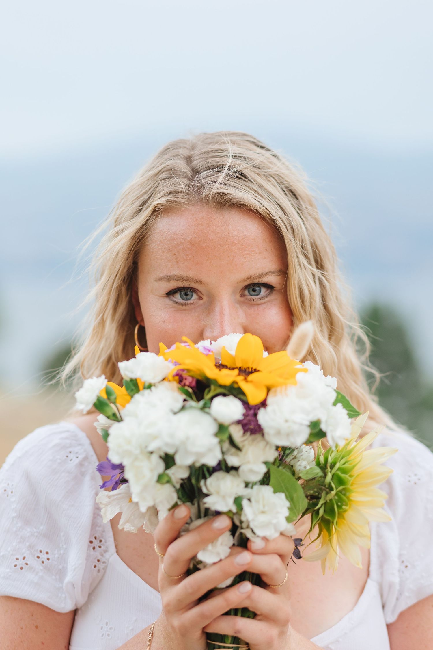 A person holds a vibrant bouquet of white and yellow flowers in front of their face against a soft blue sky background.