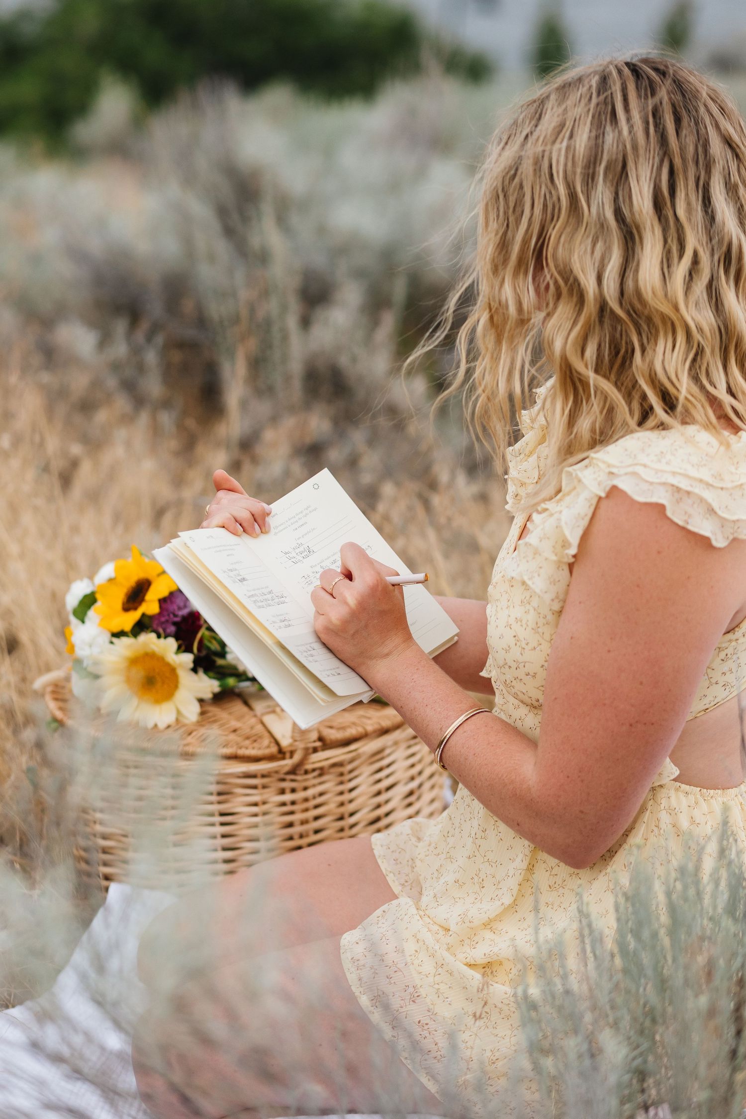 A person in a white dress reads a book while sitting in a field with sunflowers and a wicker basket.