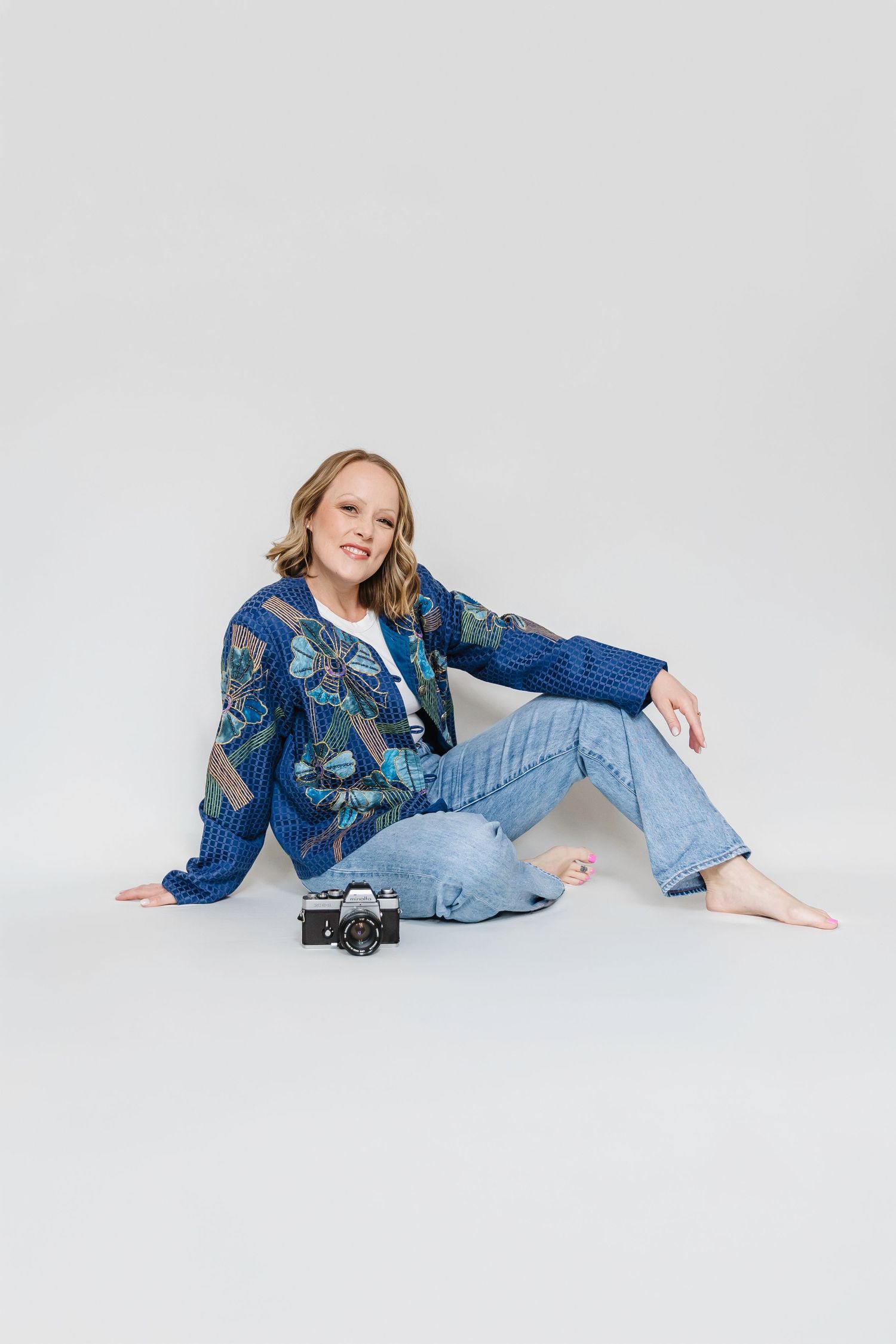 Woman in blue floral jacket and jeans sitting on floor with vintage film camera, smiling at camera.