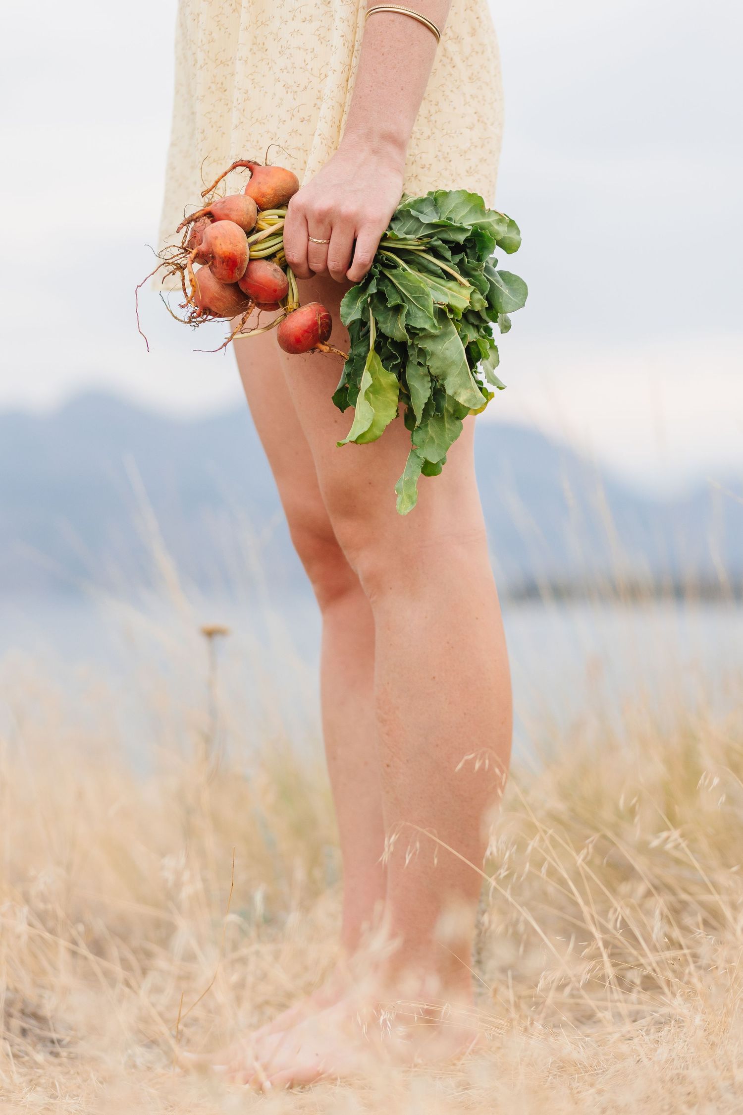Bare legs and feet stand in a grassy field while holding fresh carrots with green tops against a mountain backdrop.