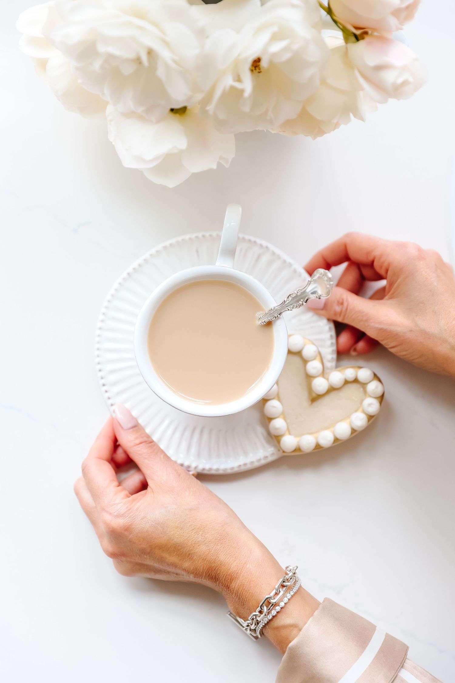 Overhead view of coffee cup with heart-shaped cookie and white peonies on white surface.