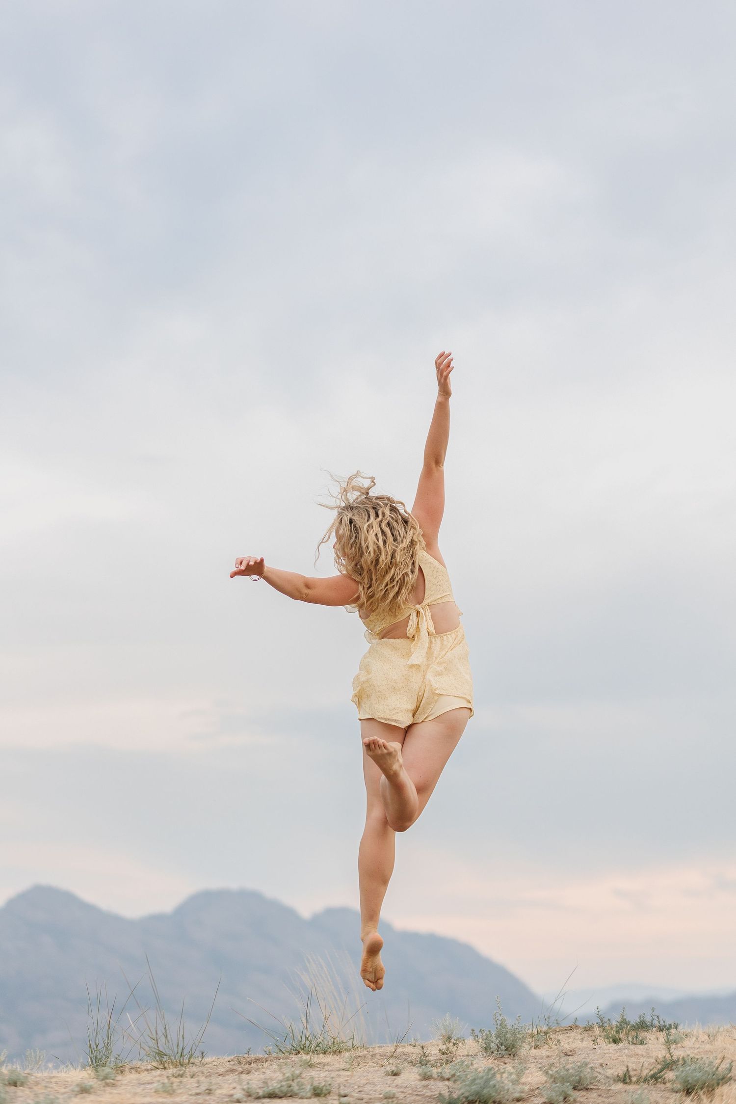 A figure in light clothing leaps joyfully against a cloudy sky with mountains visible in the background.