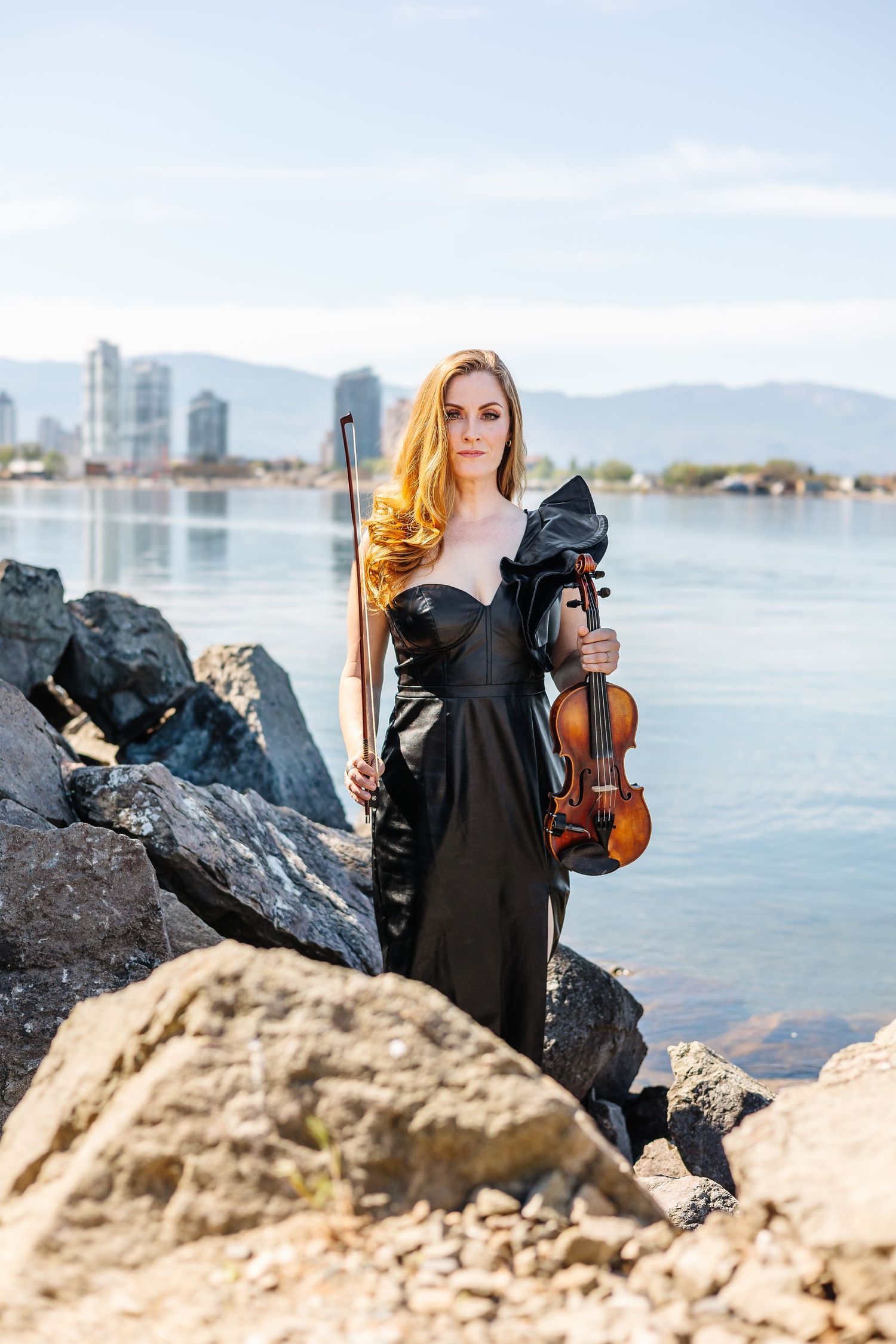 Violinist in black dress posing among coastal rocks with city skyline in background.