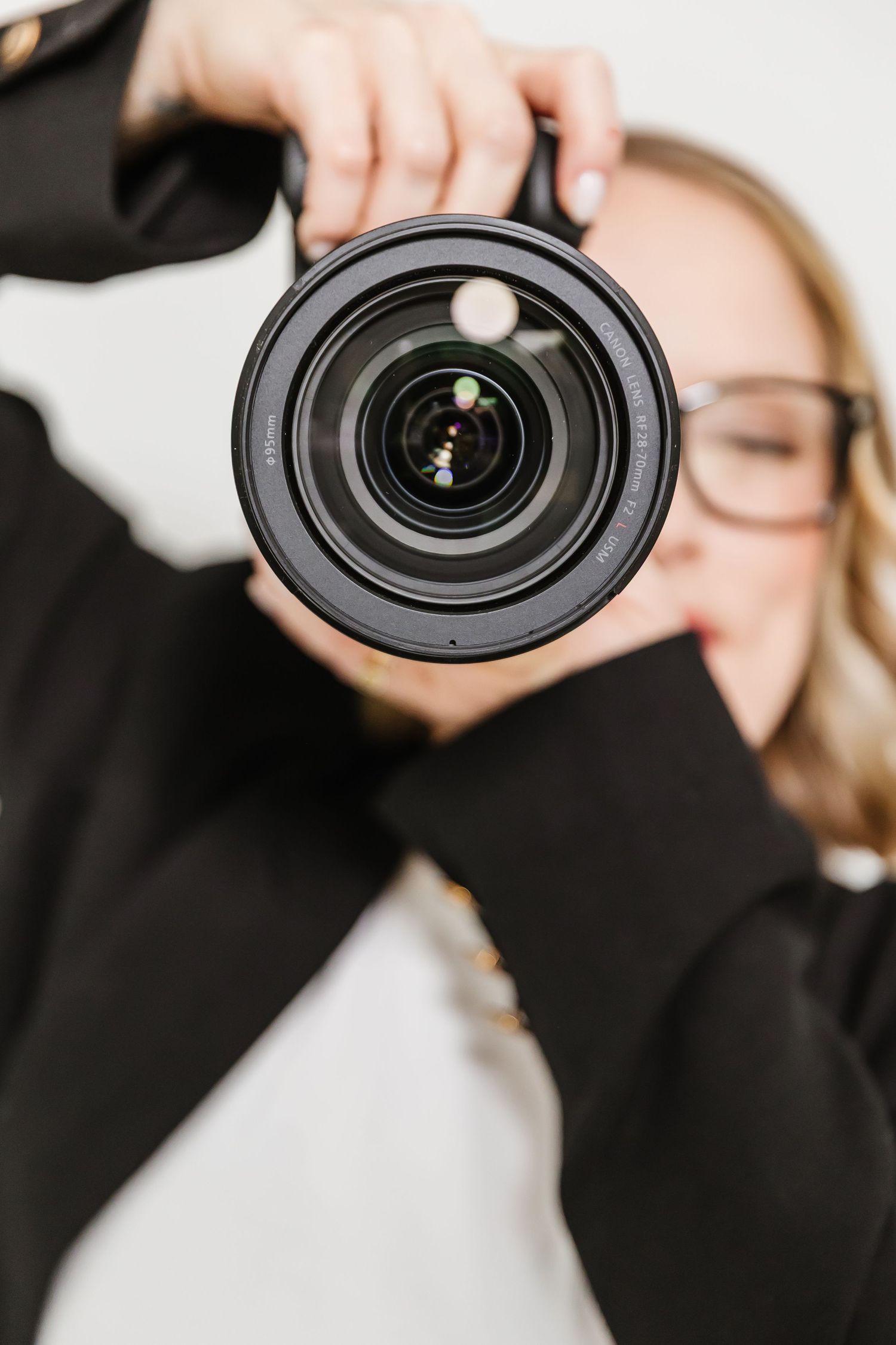 Blonde woman in black jacket holding a DSLR camera lens toward viewer, wearing glasses, on white background.