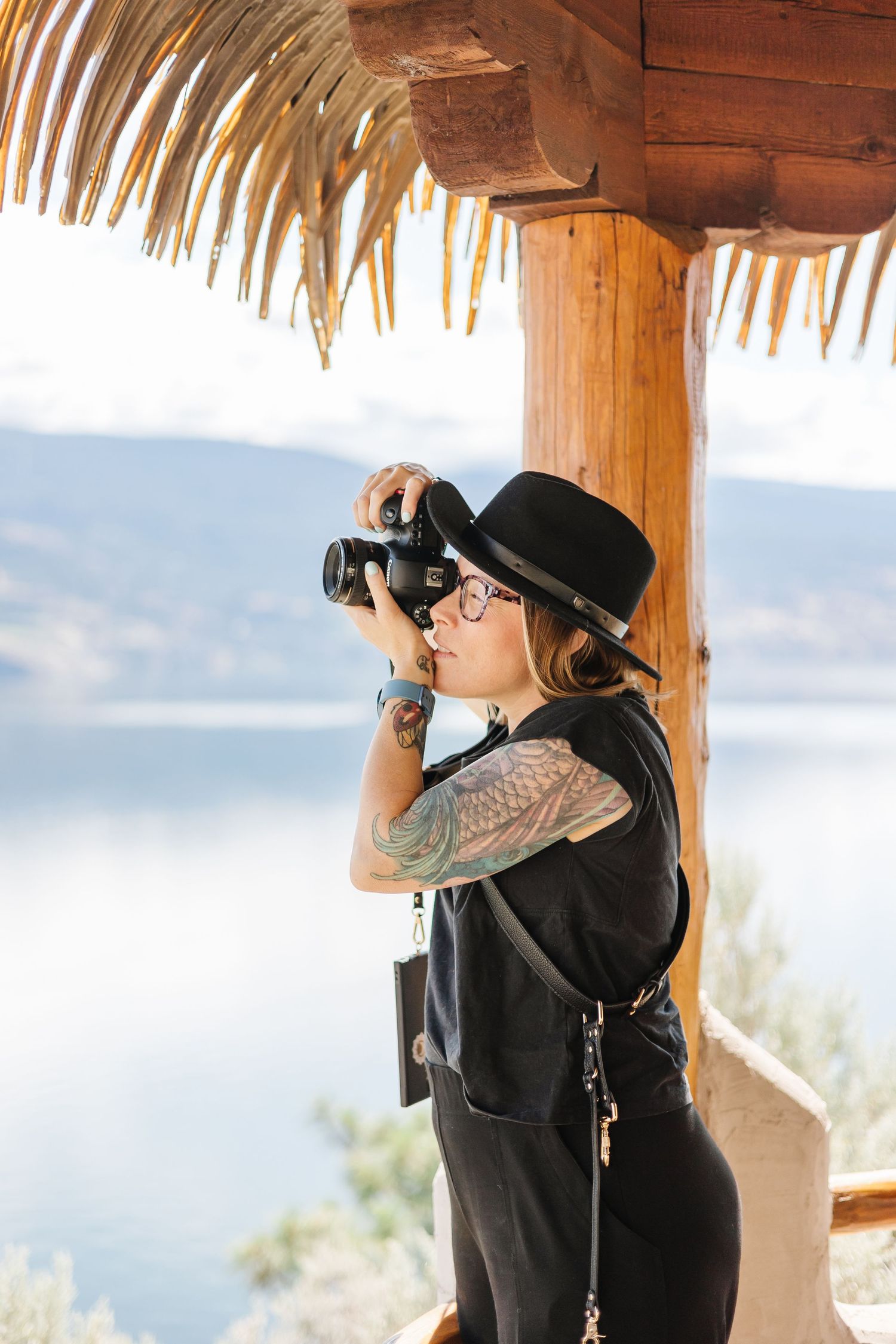 A photographer in black attire and hat takes photos from a covered porch with palm fronds overhead.