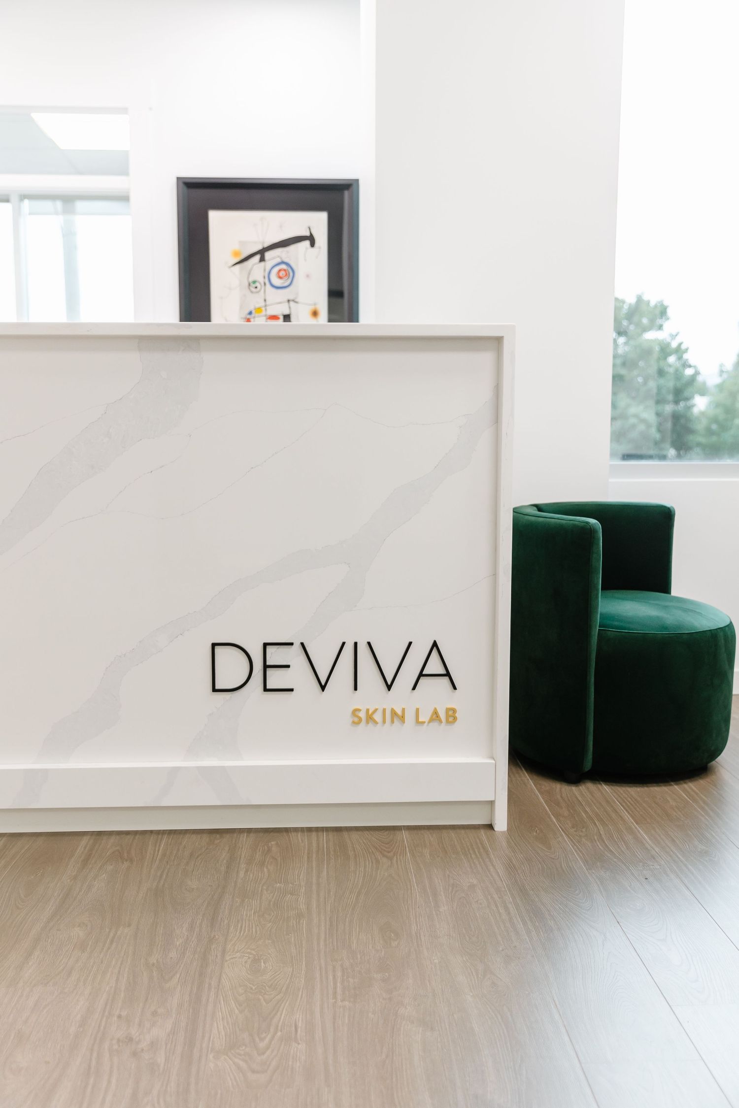 Modern medical spa reception area with white marble desk, emerald green velvet ottomans and light wood flooring.