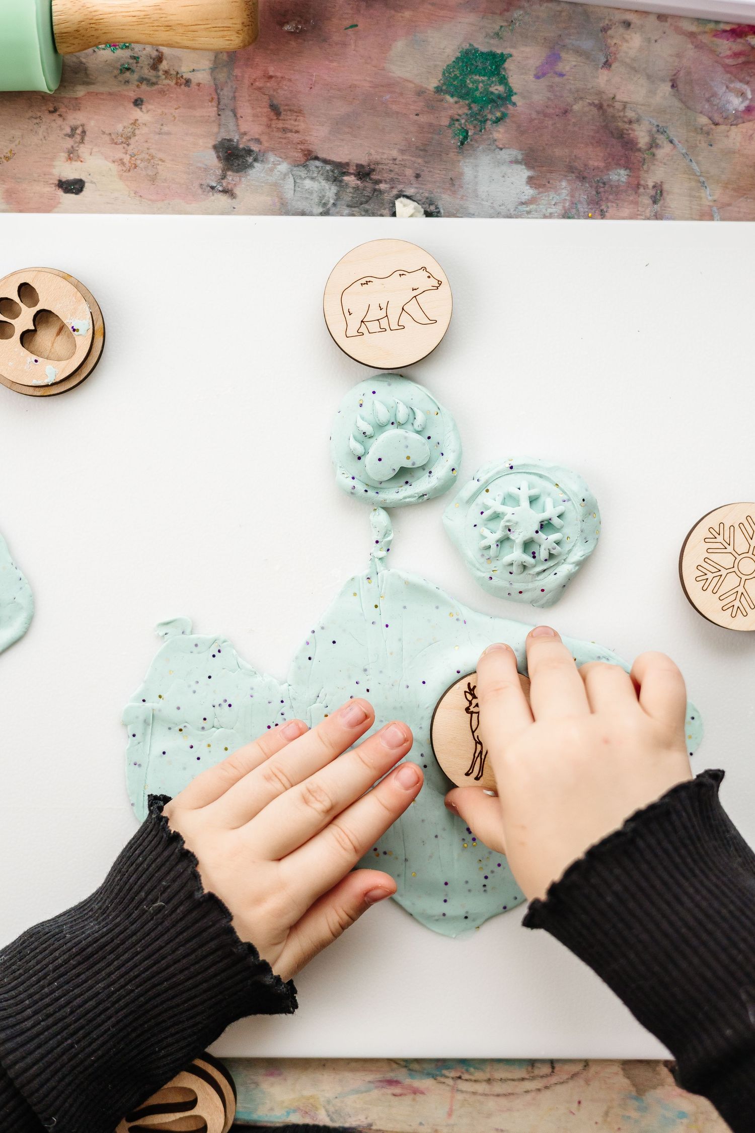 Hands pressing and shaping light blue clay or dough on a white surface with wooden stamps nearby.