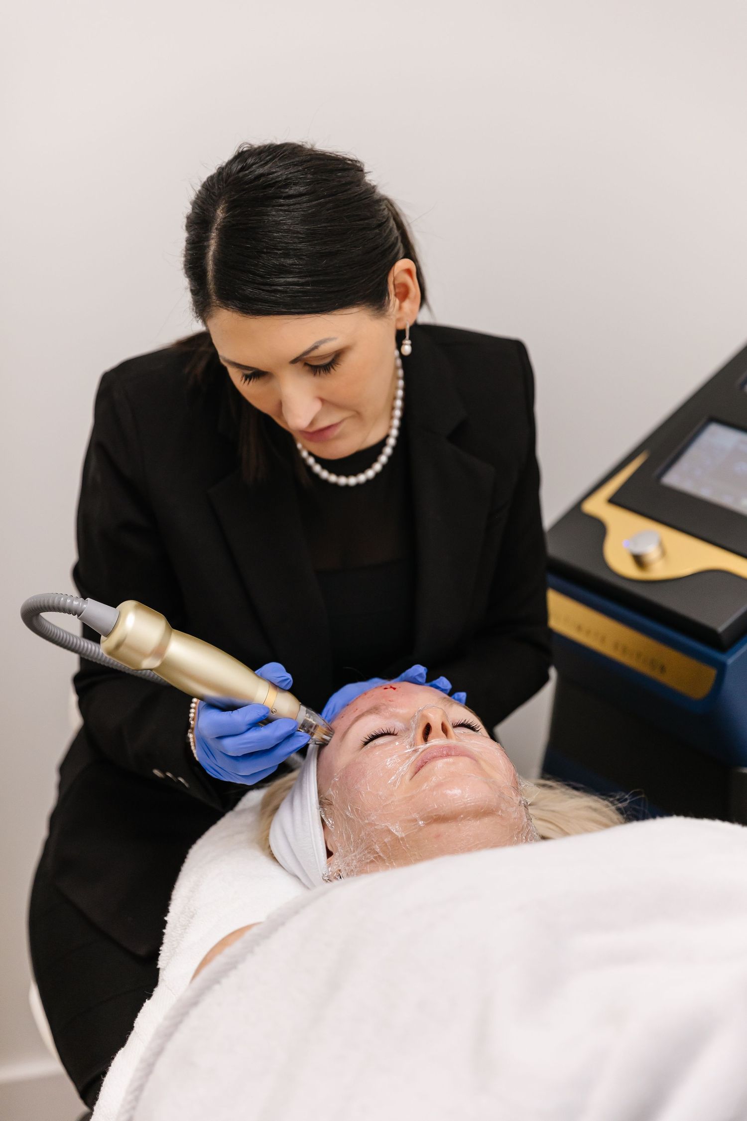 Professional esthetician performing a facial treatment on client in spa using blue medical gloves and black attire.