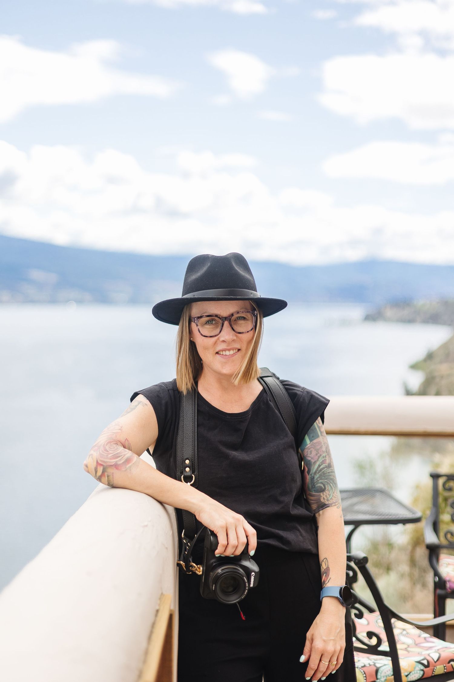 Photographer with a camera wearing a black hat and glasses stands by a railing overlooking a scenic mountain lake view.