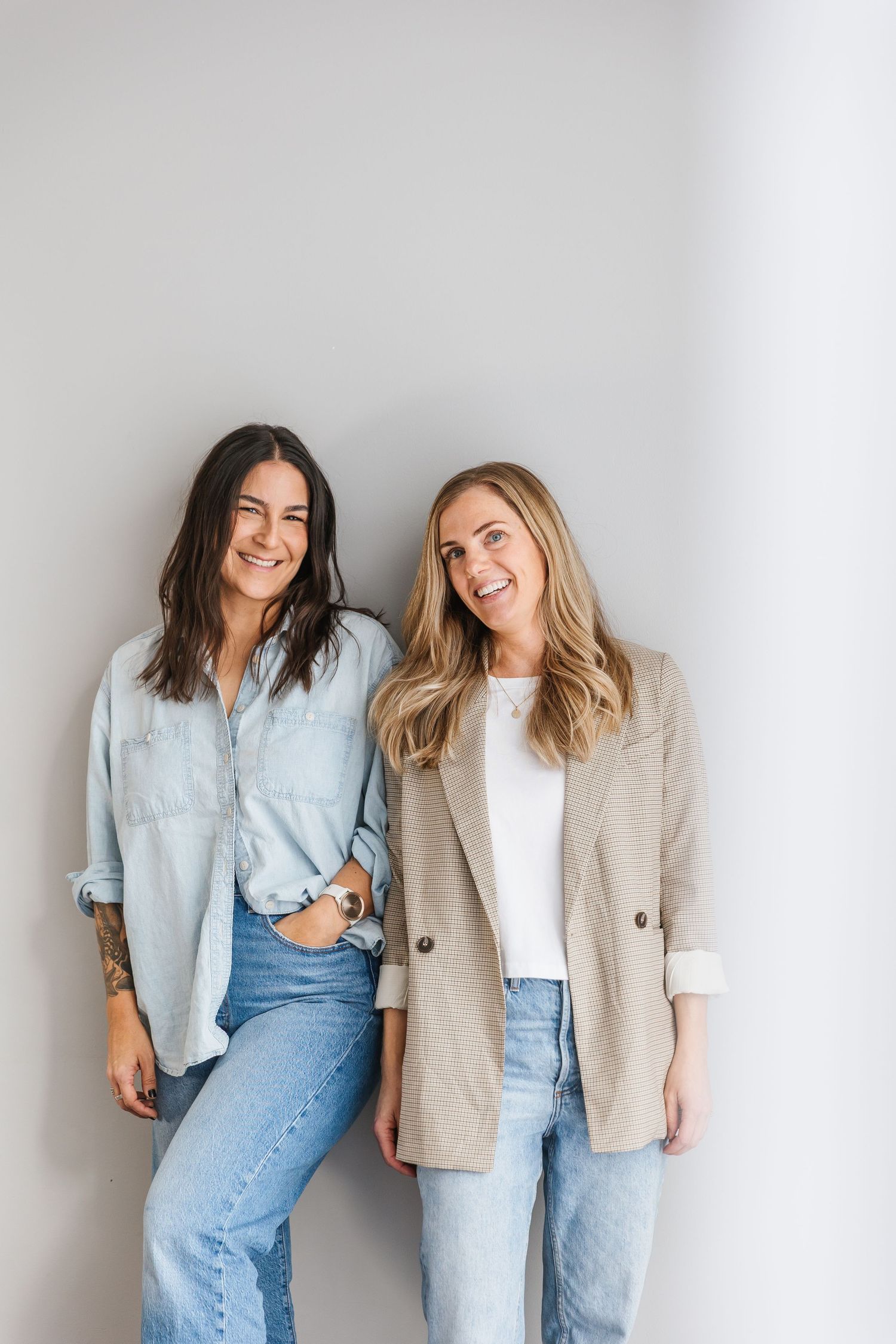 Two friends in casual denim and neutral outfits lean against white wall while laughing together.