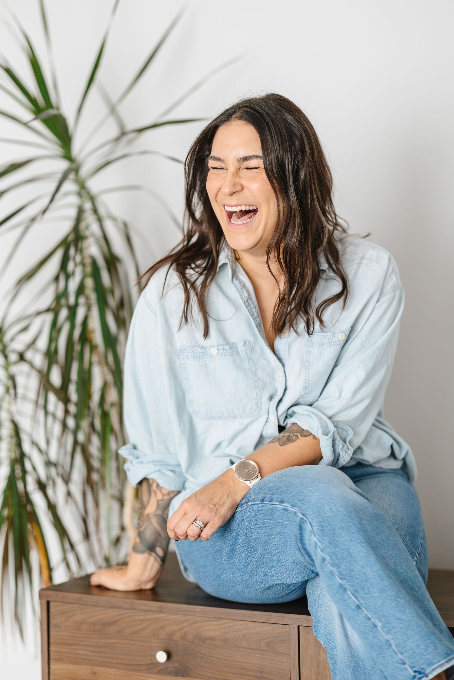 Person in light blue shirt and jeans sits casually on wooden furniture while laughing against white wall with plant.