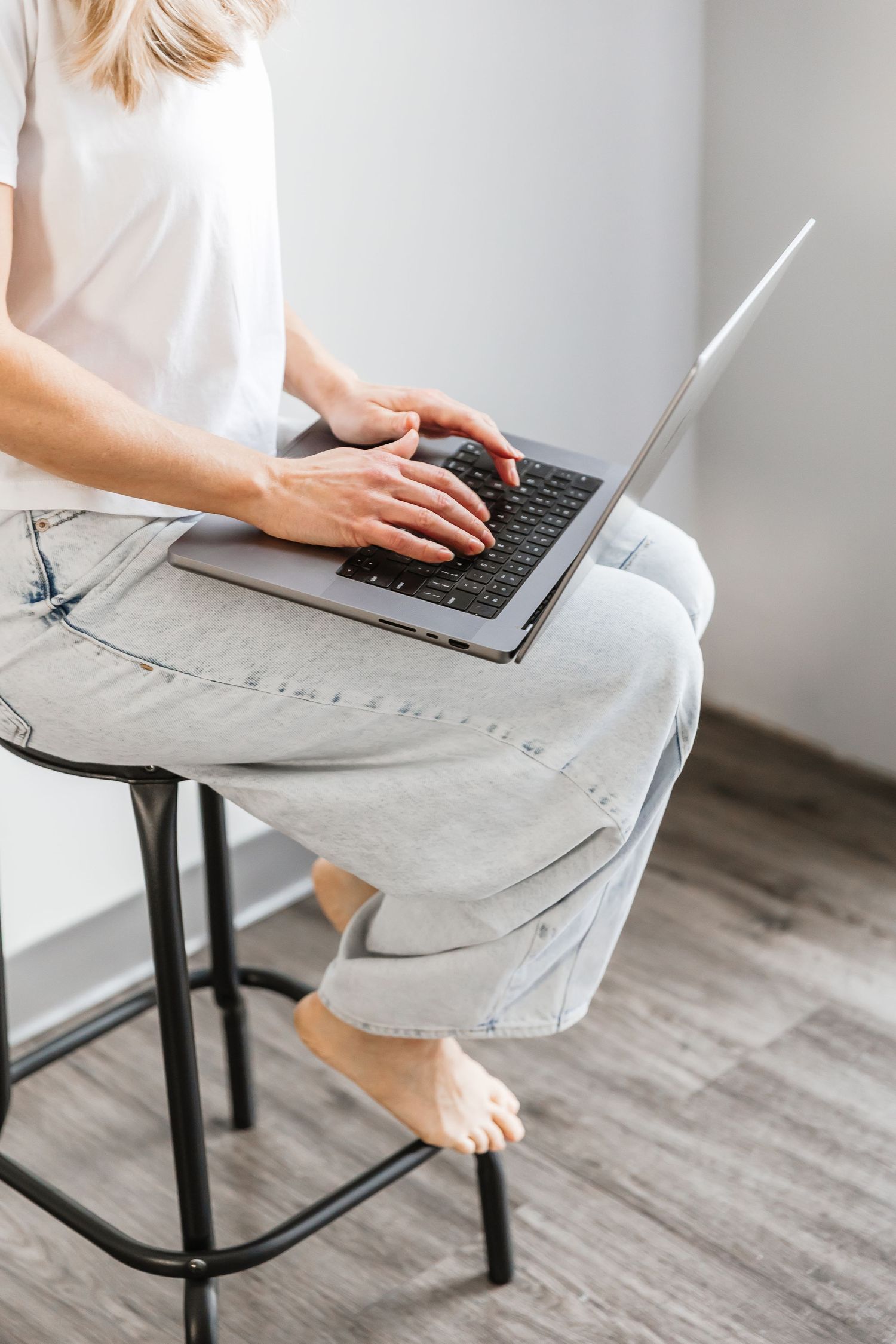 Person in white shirt and gray pants typing on laptop while sitting on black stool with bare feet.
