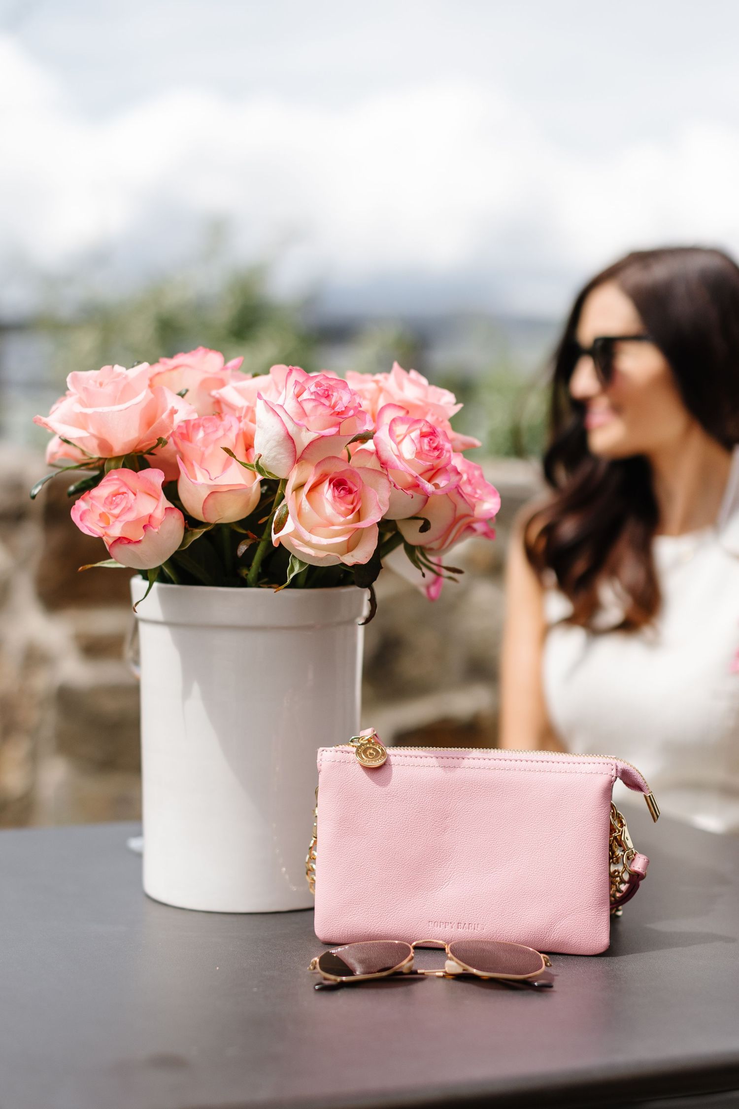 White vase of pink roses with pink wallet on outdoor table setting.