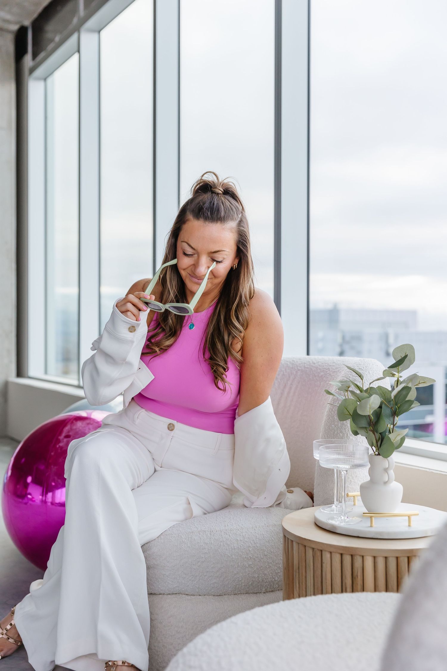 Woman in pink top and white outfit relaxing on modern couch by large windows.
