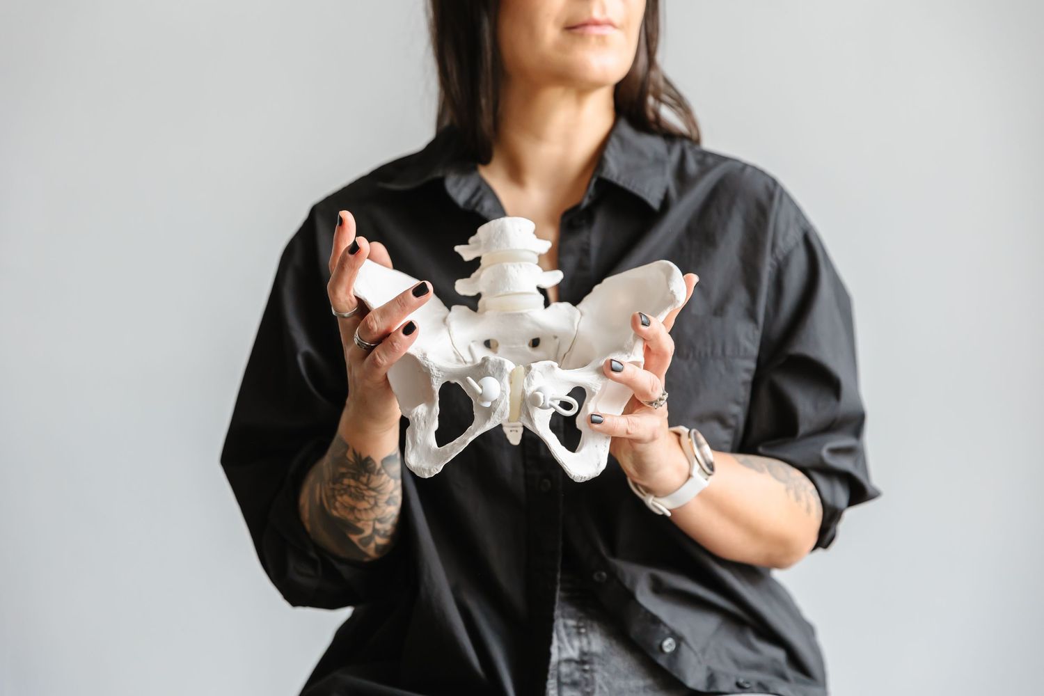 Hands with tattoos hold a white anatomical model of a human pelvis against a gray background.