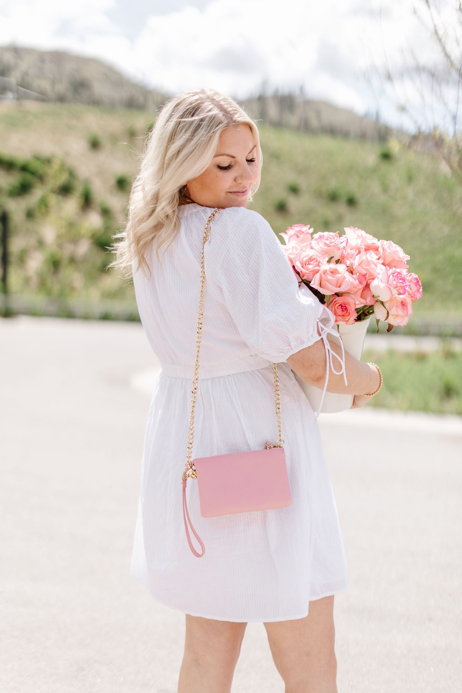 Person in white outfit carrying pink purse and roses against nature backdrop.
