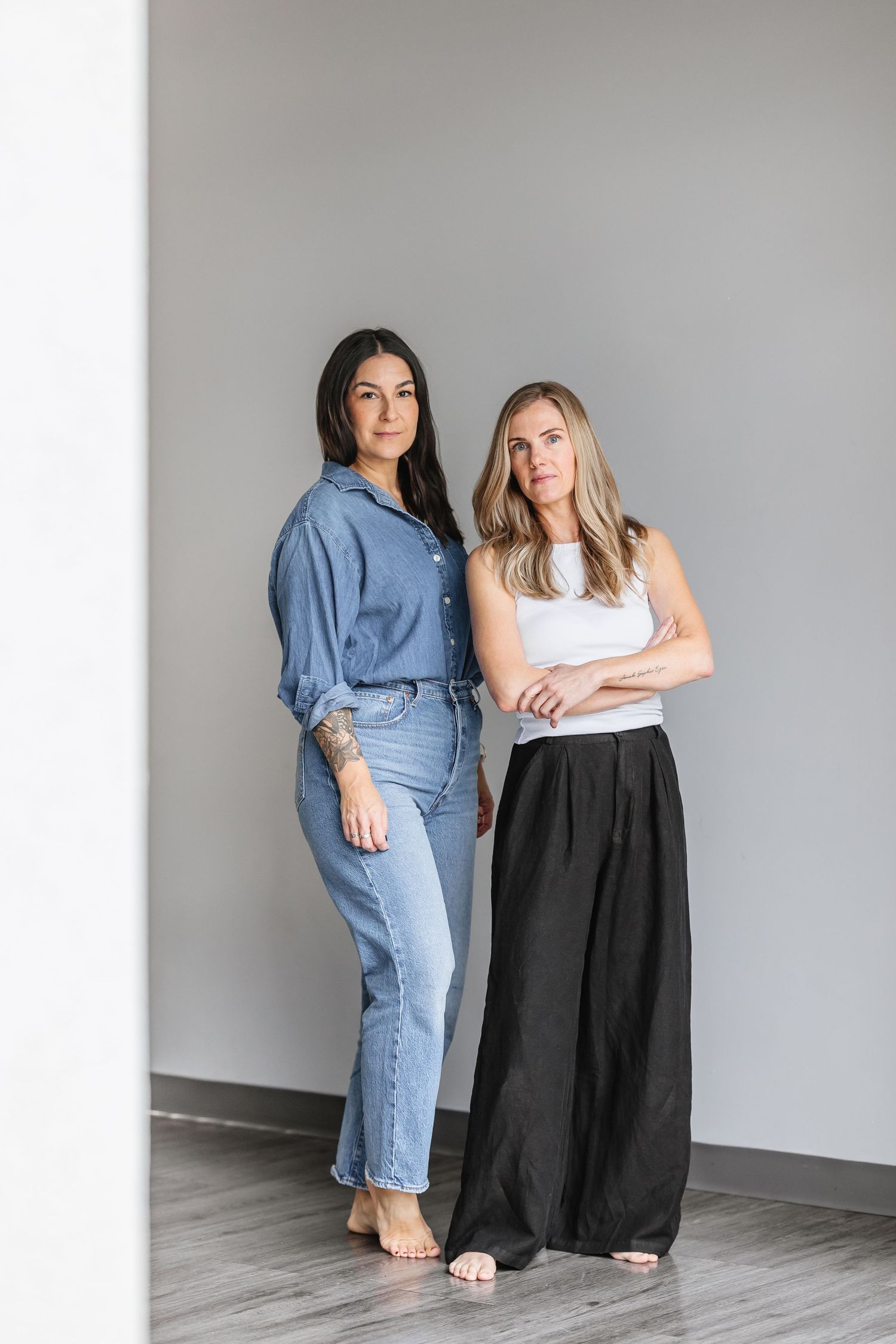 Two people in casual outfits pose together against a white wall in a minimalist studio setting.