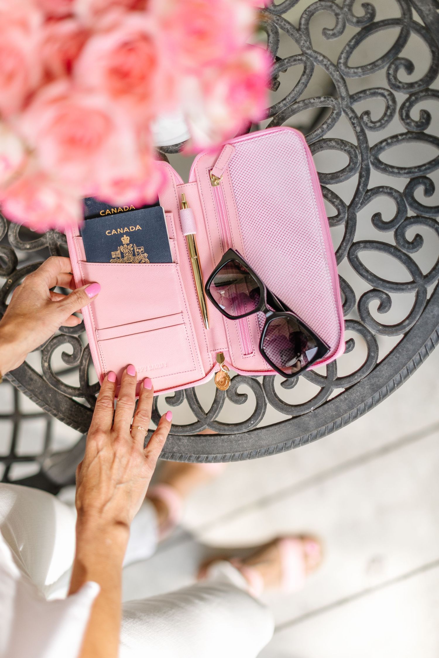 Hands displaying pink passport holder with sunglasses on decorative metal table.