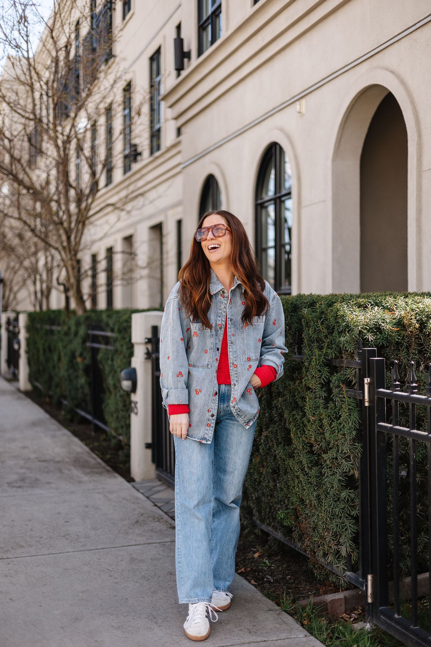 Woman in denim jacket with heart embroidery, red sweater, wide-leg jeans, and white sneakers laughing outdoors.