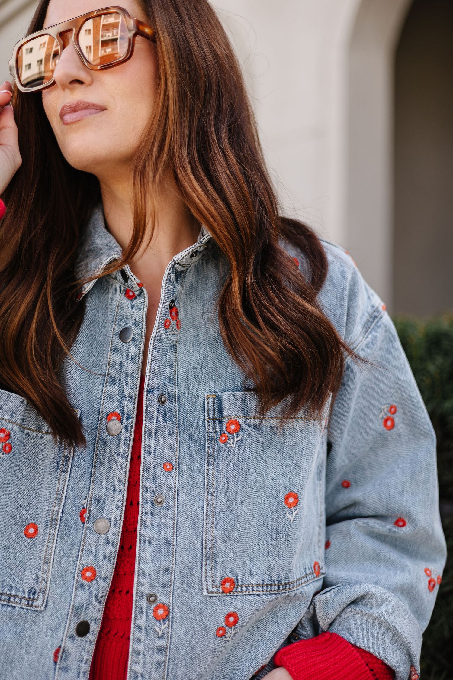Woman wearing a floral embroidered denim jacket with red flowers, paired with a red top and tortoiseshell sunglasses.