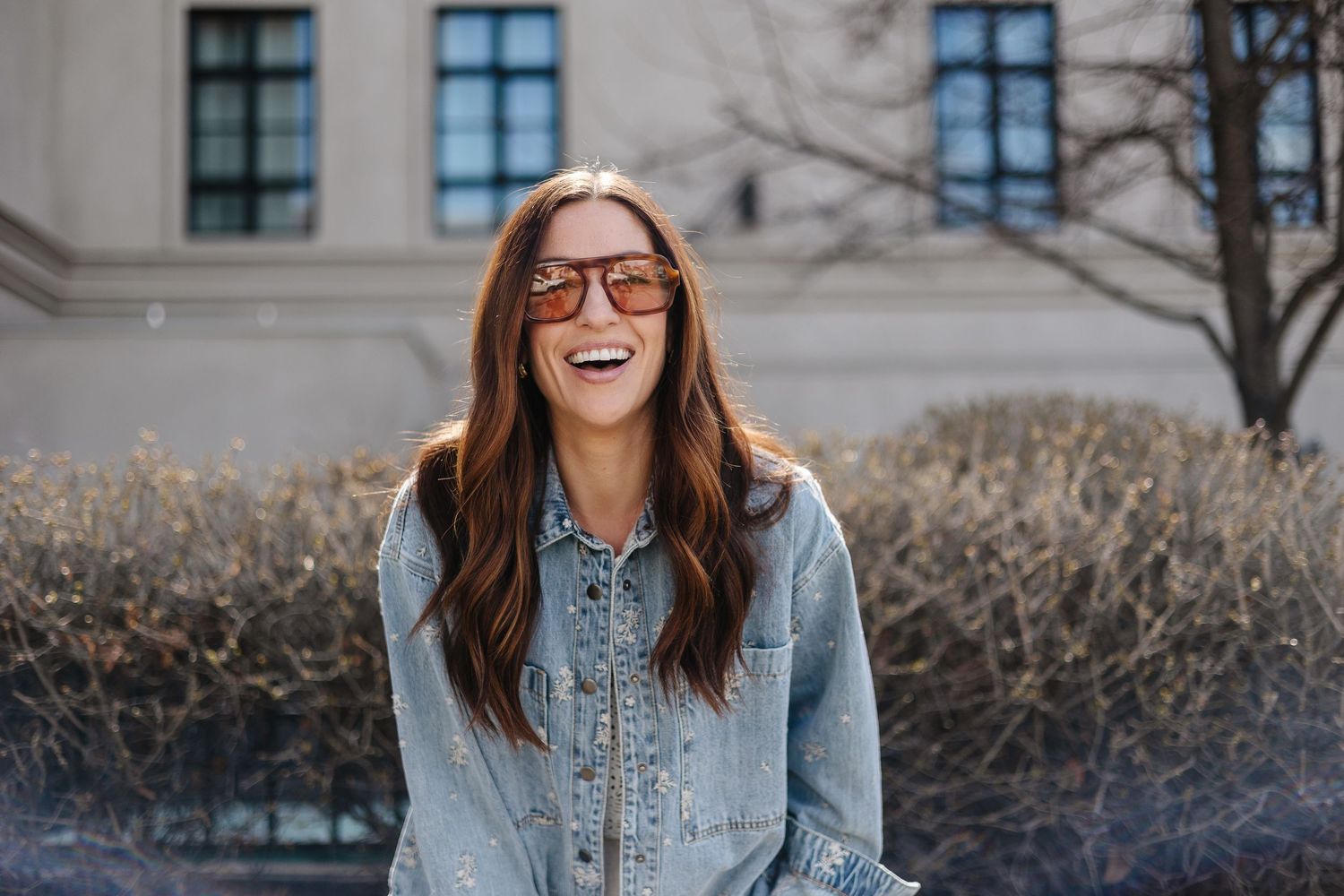 Smiling woman with long brown hair wearing sunglasses and a denim jacket outdoors near shrubs.