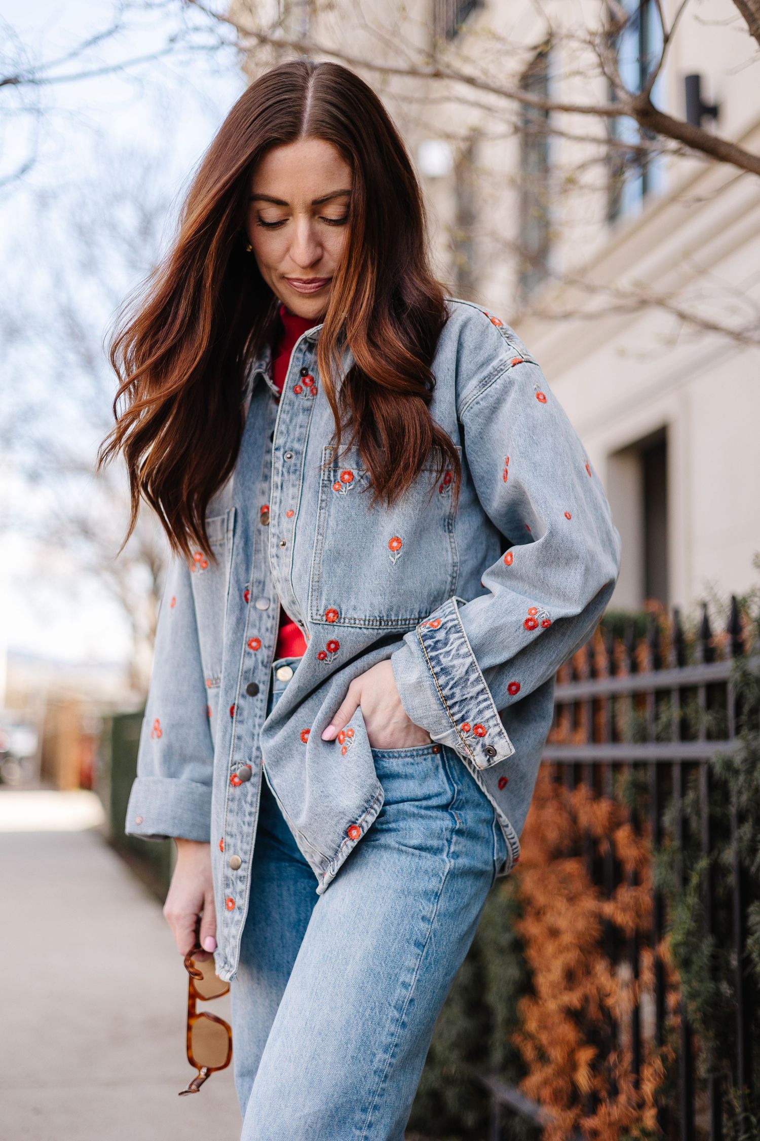 Woman in denim jacket and jeans with red floral embroidery, holding sunglasses on a city street.