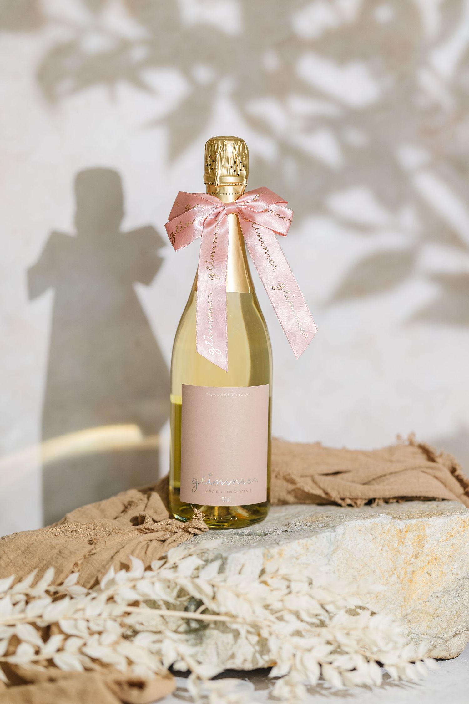 A small glass bottle decorated with a delicate pink bow sits on a textured beige surface in soft lighting.