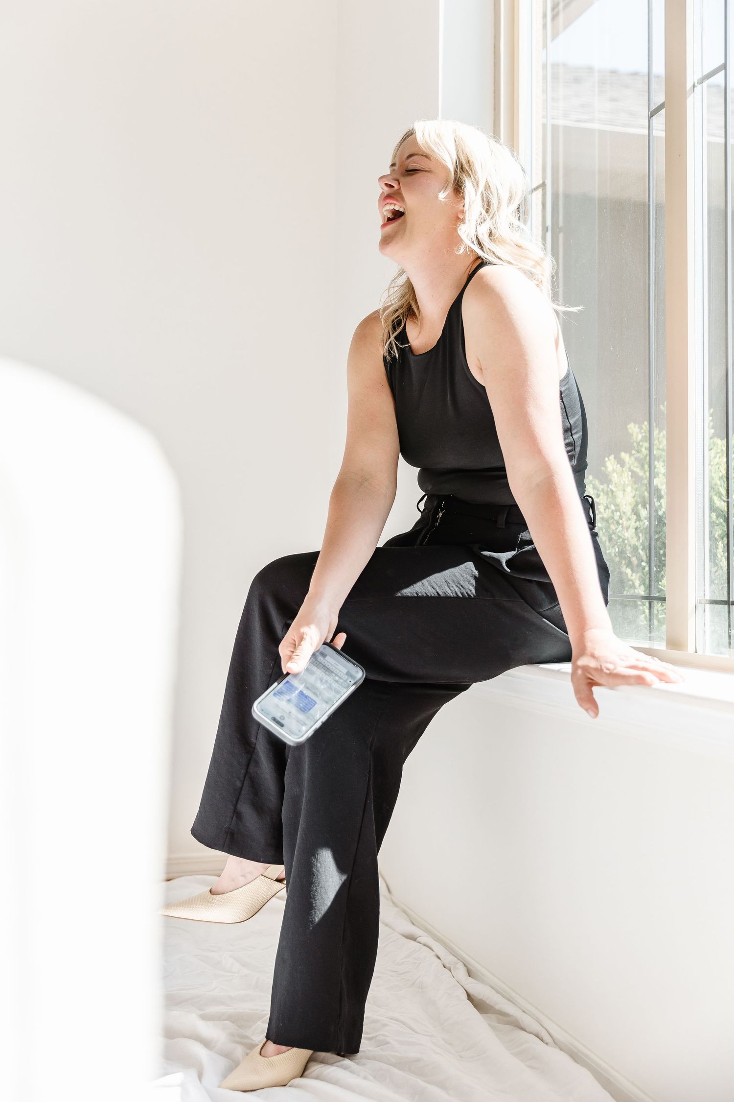 Woman in black dress sitting elegantly by window with natural light streaming in.