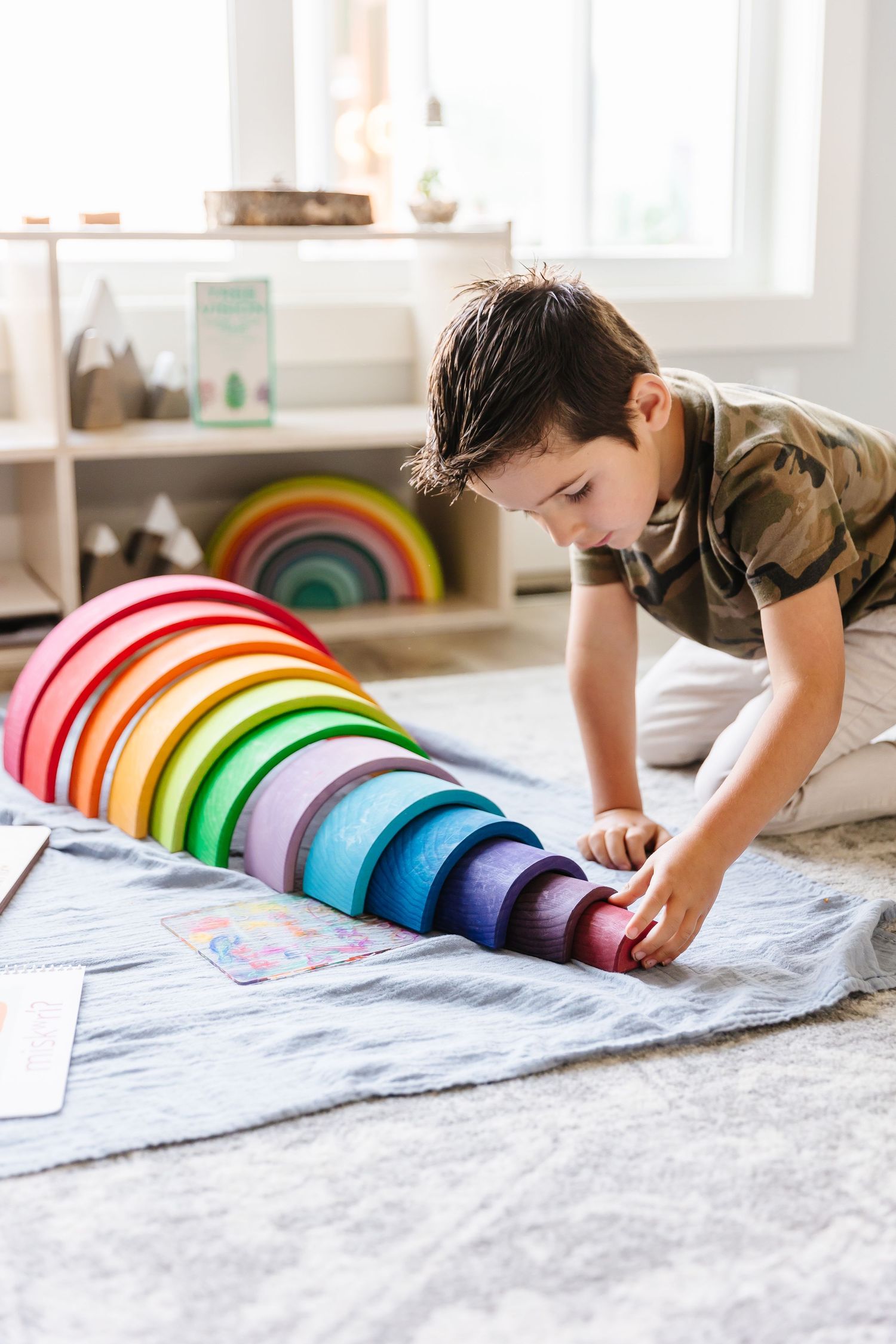 Rainbow-colored wooden arch toy arranged on light gray blanket in playroom setting.