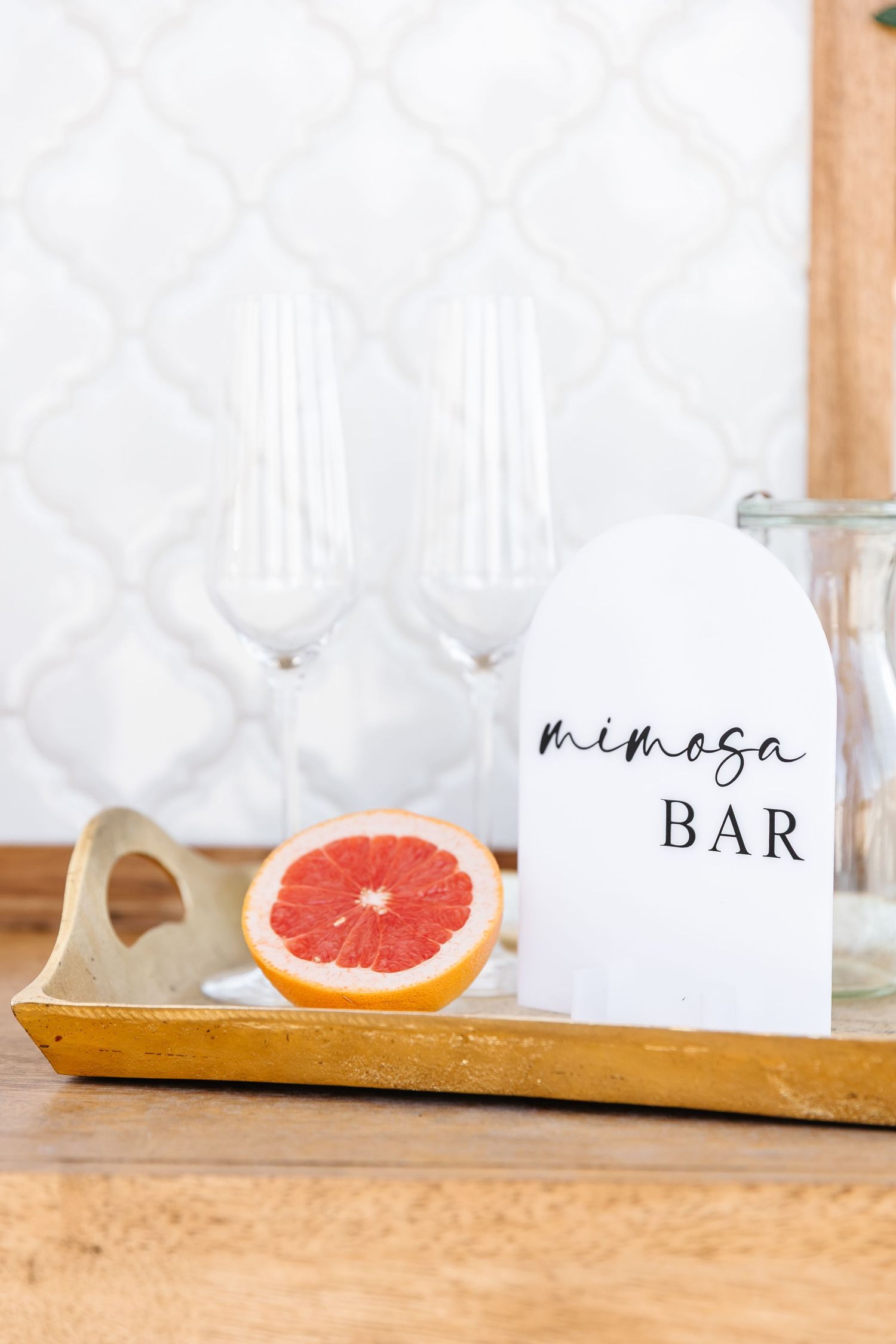 A mimosa bar setup with champagne flutes, fresh grapefruit, and a white ceramic container labeled 'Mimosa Bar' on a wooden tray.
