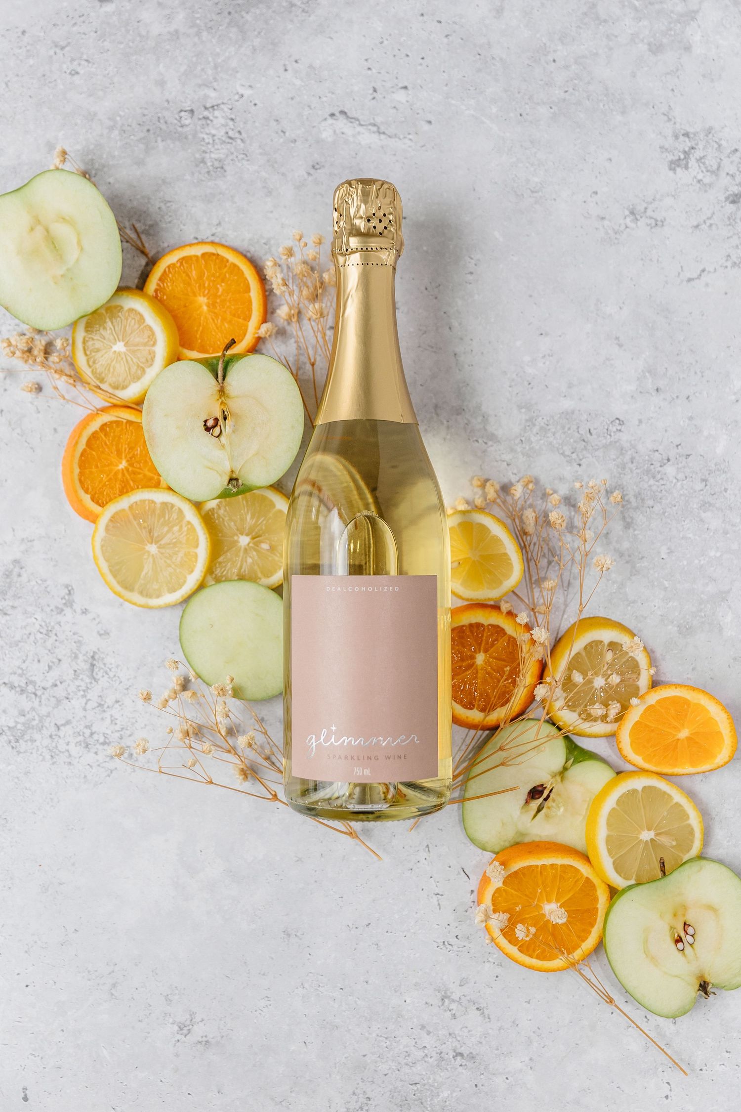 Gold champagne bottle with pink label surrounded by fresh citrus slices and green apples on light gray background.