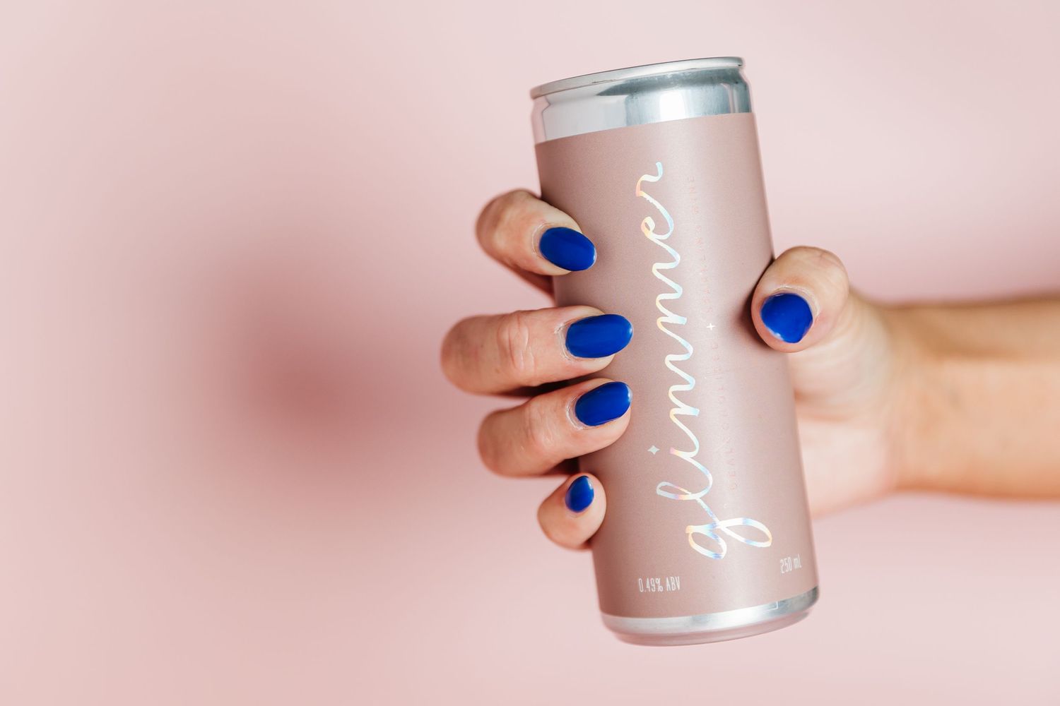 Hand with bright blue nail polish holding a silver energy drink can against a soft pink background.