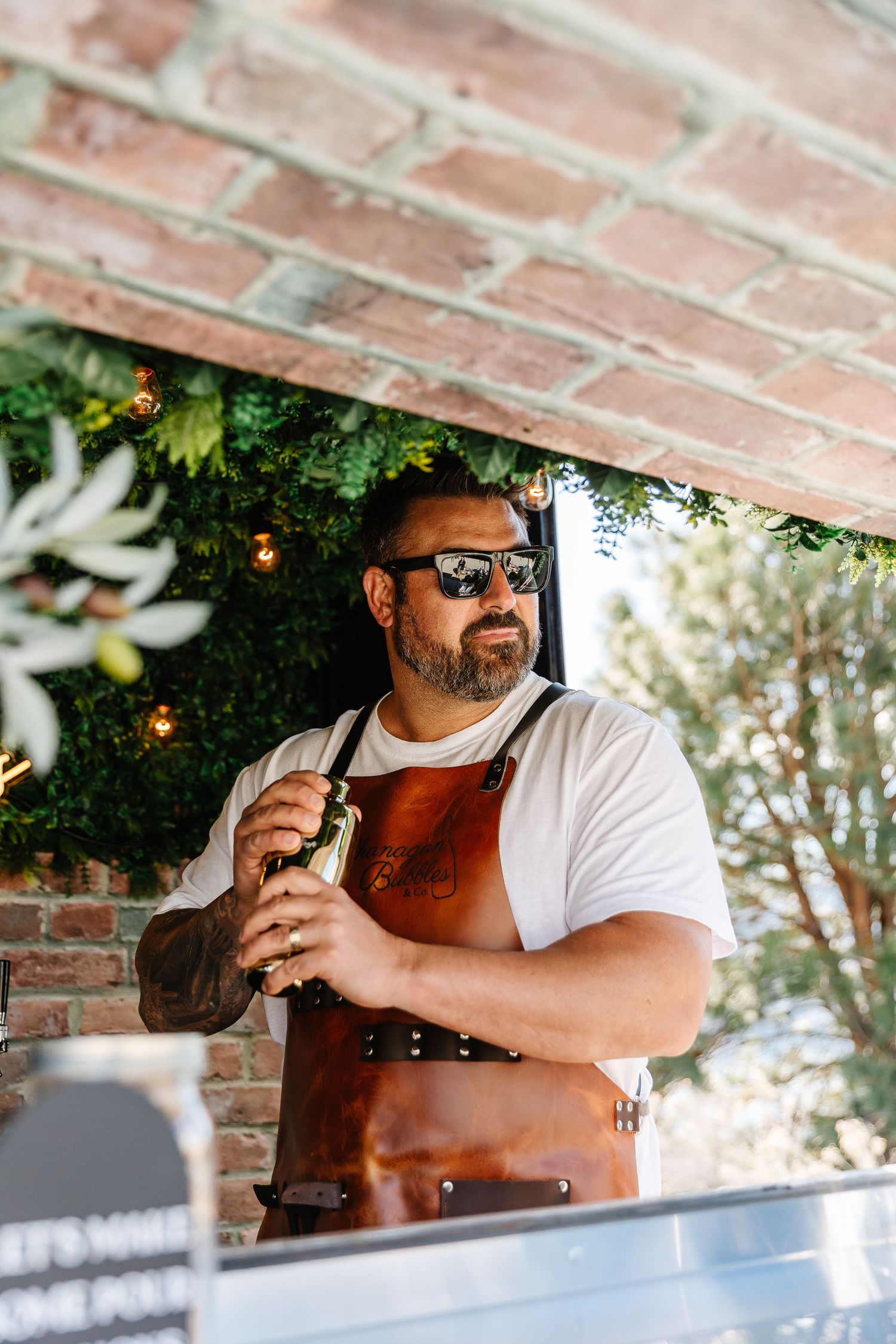 A bartender in sunglasses and white shirt works behind a counter with exposed brick wall and greenery visible.