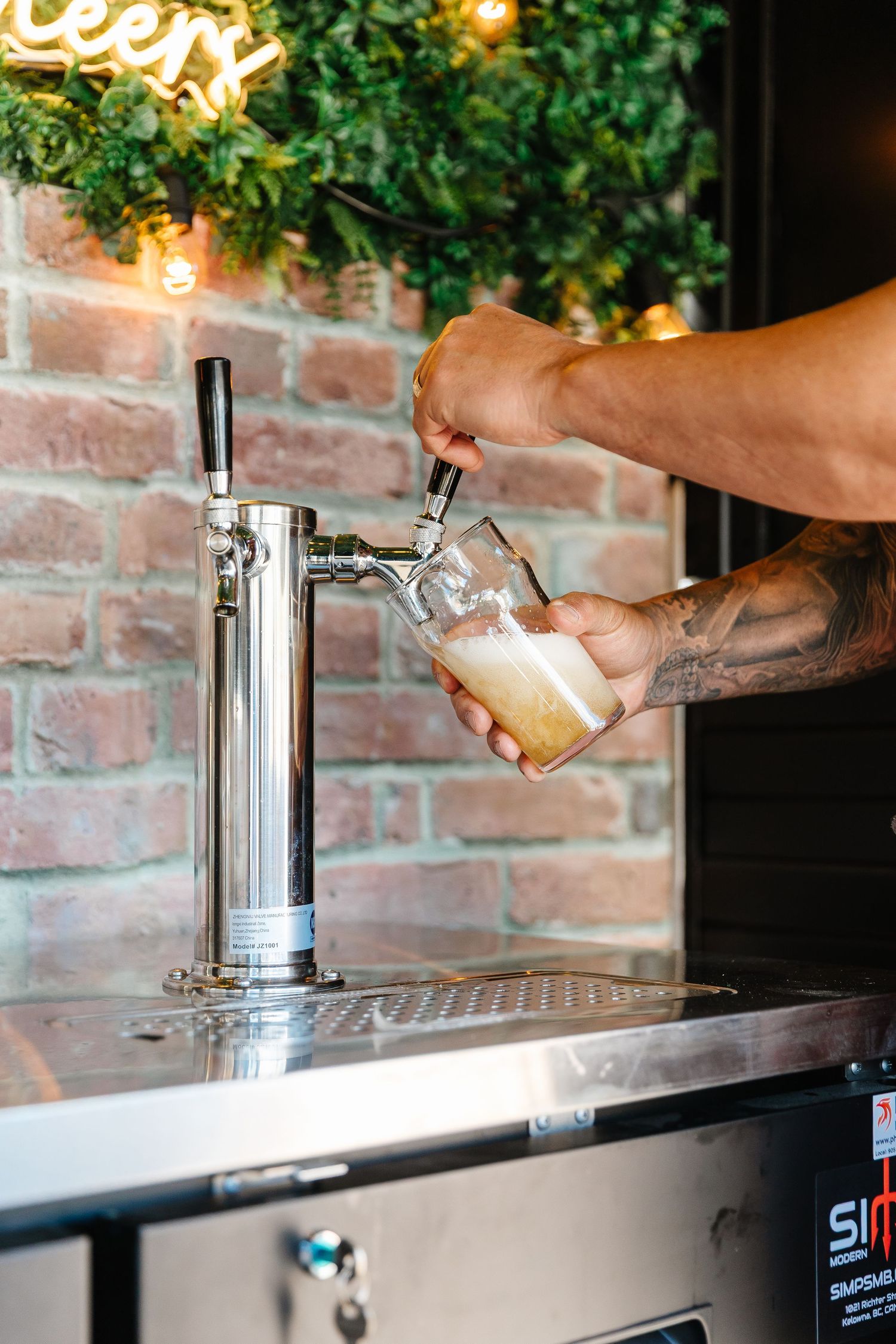 Tattooed hands pour a draft beer from a chrome tap at a brick-walled bar with ambient lighting.