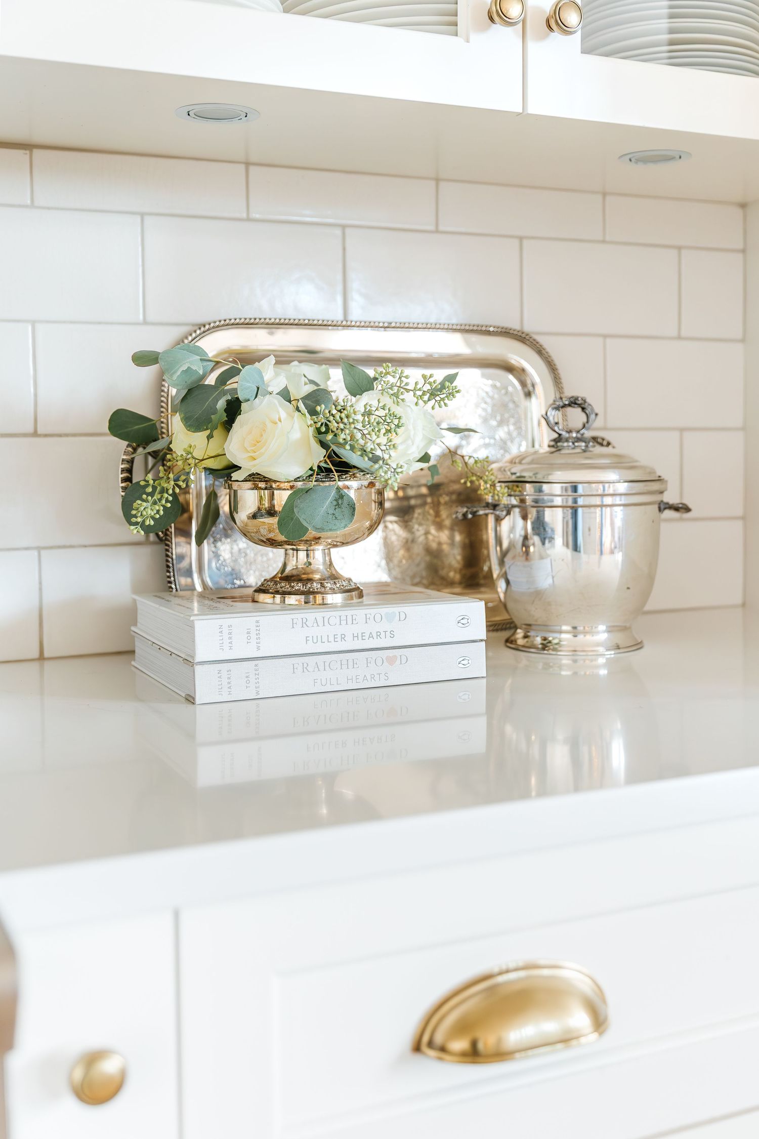 White kitchen counter styled with silver tray, floral arrangement, stacked books, and gold cabinet hardware against subway tile.