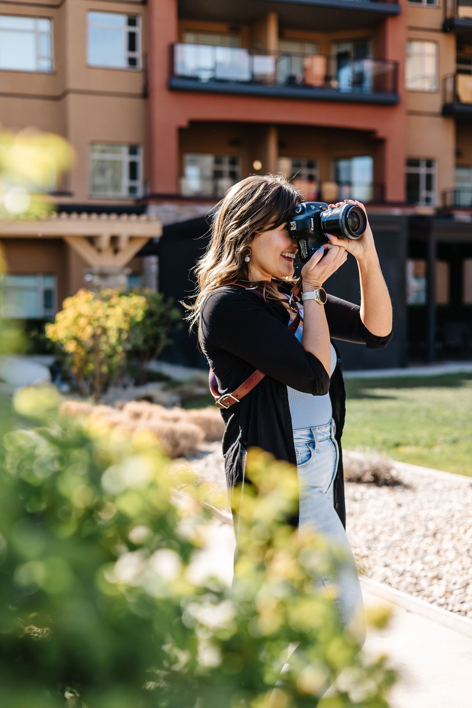 A photographer with a camera takes photos through foliage near brick buildings.