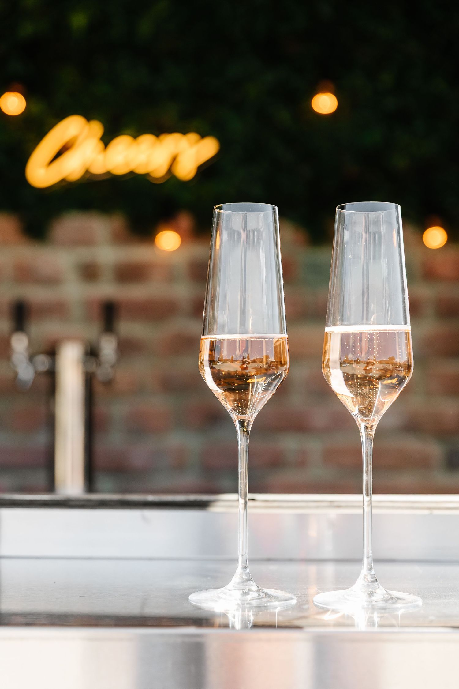 Two elegant champagne flutes filled with rose wine sit on a surface with glowing lights and a neon sign in the background.