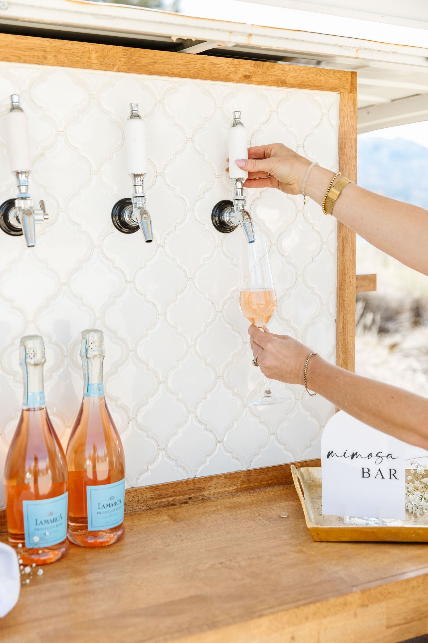Pink and orange bottles of drinks displayed on a wooden bar shelf with white decorative backdrop.