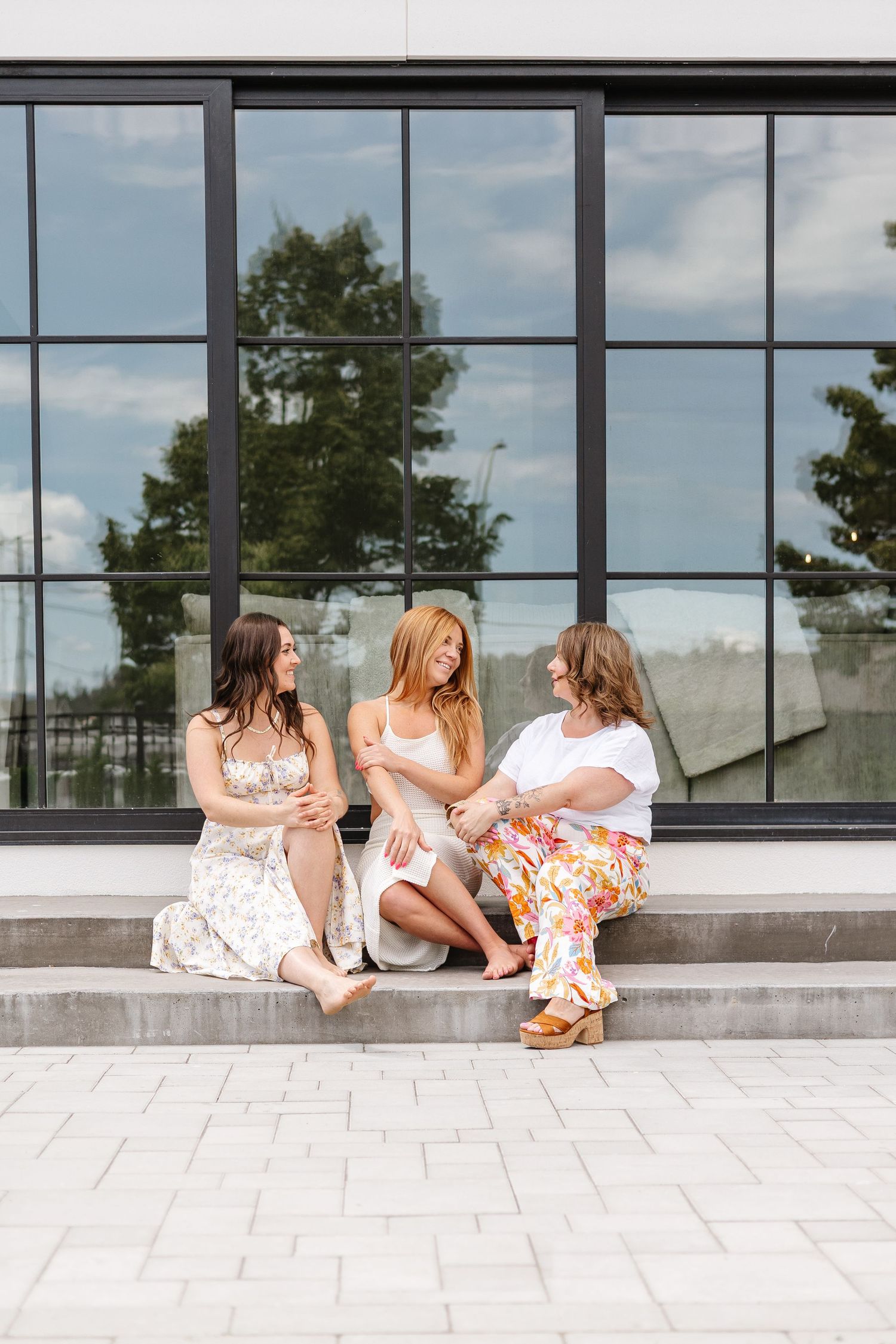 Three women in white summer dresses sit casually along a modern glass building exterior.