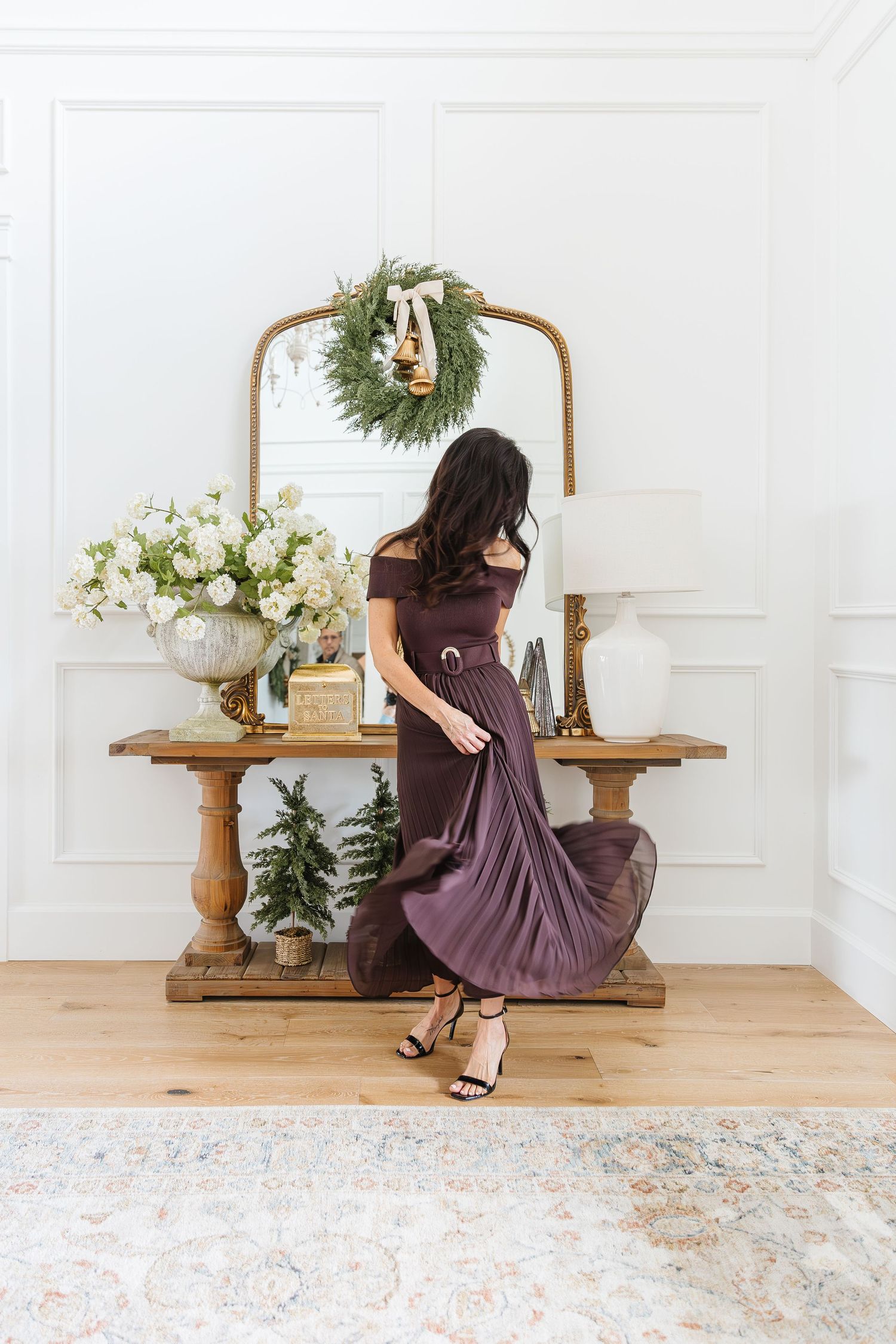 Elegant holiday scene with plum dress, ornate mirror, white floral arrangement and festive wreath in bright interior.