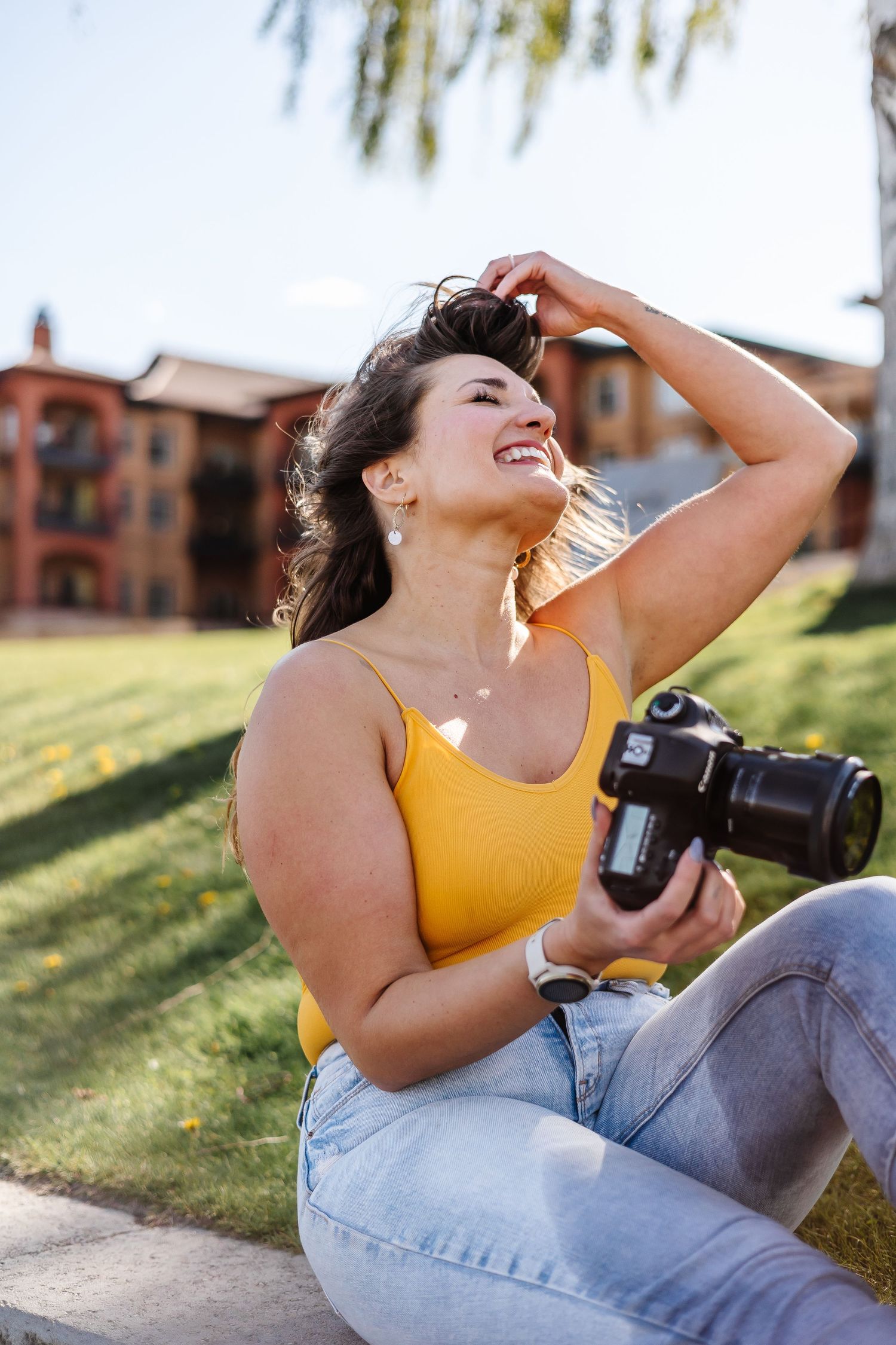 A photographer in a yellow top lies on grass with a camera near waterfront apartments.