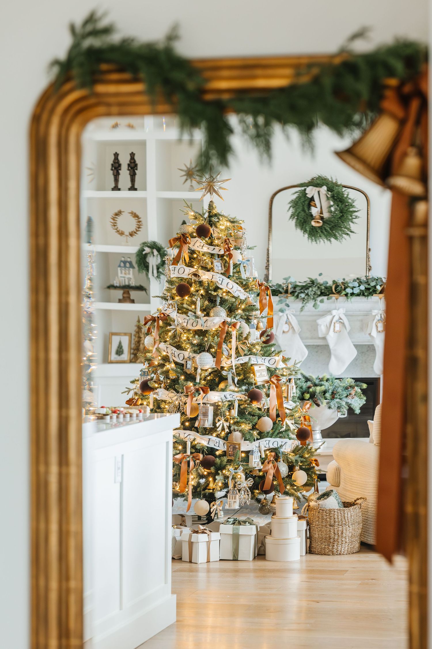Elegant Christmas living room with decorated tree, gold ornaments, gifts, fireplace garland, and wreath in white interior.