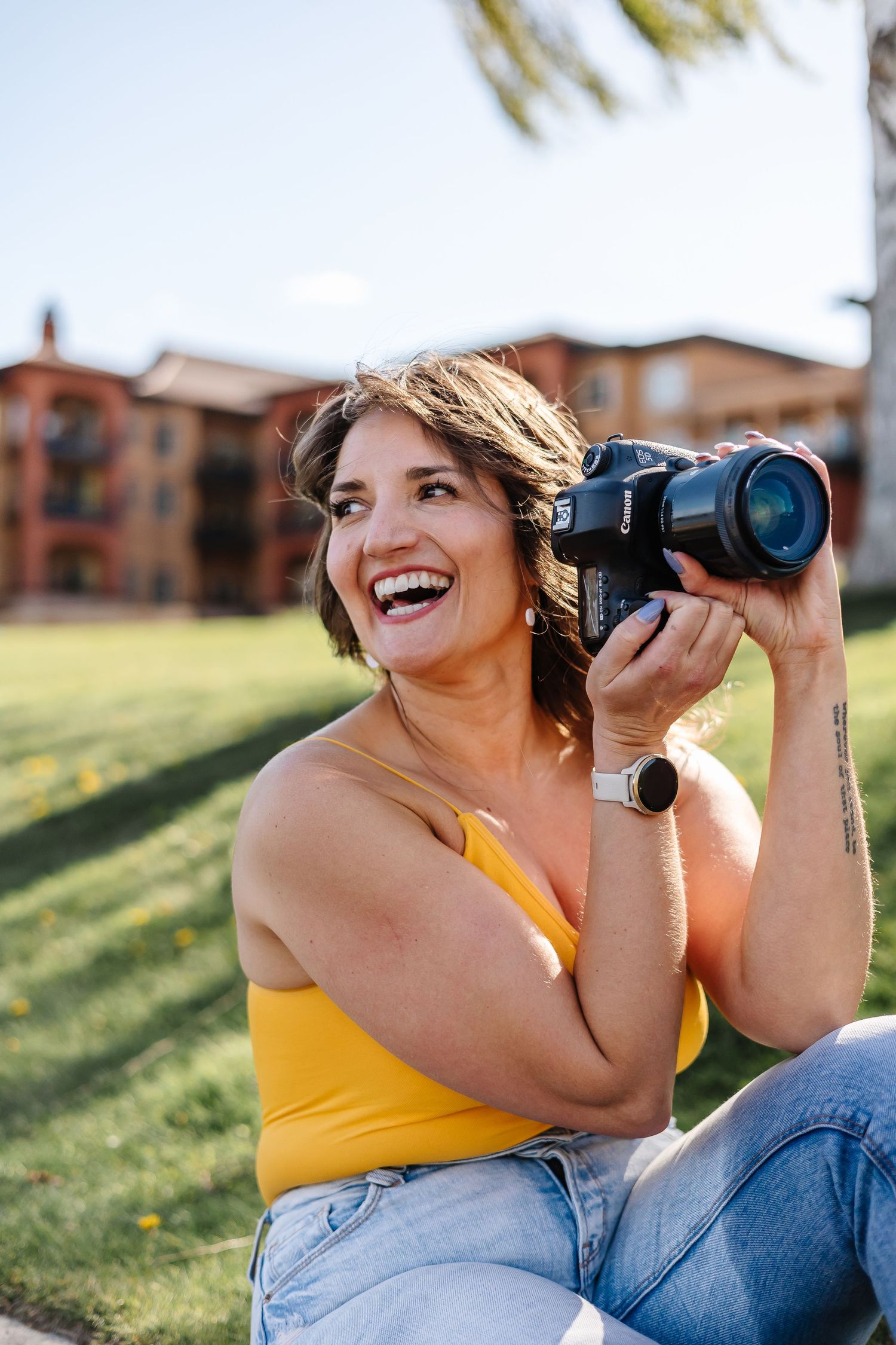 A photographer in yellow tank top and jeans laughs while holding a camera outdoors.